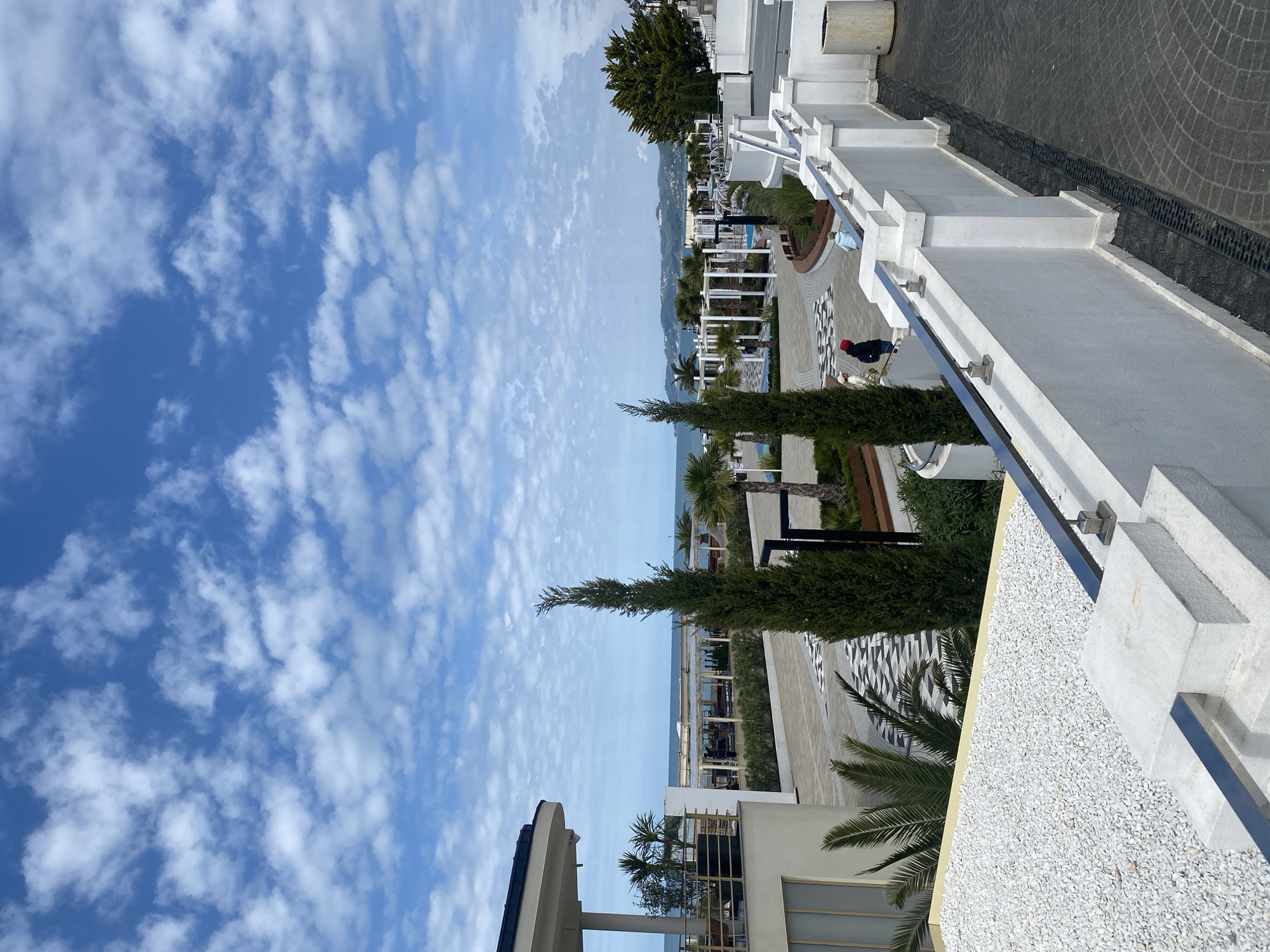 The image displays a modern resort or promenade in Sochi, Russia, overlooking the Black Sea under a partly cloudy sky. The scene is dominated by contemporary white architecture, including multi-tiered white concrete structures with minimalist railings that extend along the right side. Further back, several white pergolas and lounge areas with outdoor furniture are visible, suggesting spaces for relaxation or dining.
The landscape is meticulously designed with a mix of tall, slender evergreen trees and lush palm trees interspersed throughout. Ground surfaces vary, featuring patterned grey cobblestones, black and white geometric paving, and areas covered with bright white gravel.
Two individuals are present in the middle ground. One person, wearing a dark jacket and a red hat, stands near a low white wall or bench. Another person in lighter clothing is visible further back near a light blue structure. They appear to be casually observing the surroundings, indicative of a leisurely activity.
The time of day is daytime, with soft lighting and a blue sky dotted with scattered white clouds, suggesting pleasant weather conditions. The calm sea stretches towards the horizon, completing the coastal resort setting. The overall mood conveyed is one of tranquility and modern elegance. No visible text is present in the image.