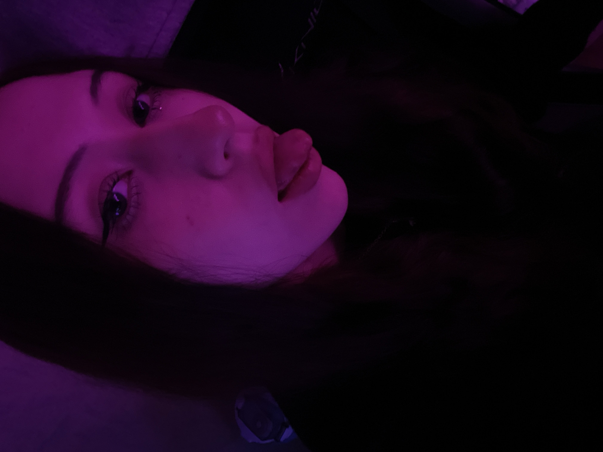 The image presents a close-up portrait of an individual, likely female, with dark hair, illuminated primarily by a strong magenta-purple light source positioned to the viewer's right. The subject's face is angled slightly to the right, maintaining direct eye contact with the camera. Both eyes feature prominent black winged eyeliner, and a small reflective spot or tear is visible below the subject's right eye (viewer's left). The mouth is open with the tip of the tongue extended. A faint, thin metal chain is discernible around the neck area on the right side. On the far left, a portion of an ear or a white earbud/device is visible against the dark background. The background is largely obscured by darkness, with subtle hints of purple ambient light on the left and a faint white/silver text or logo, appearing to read "K/NG" (suggesting a brand or 'gaming'), on a dark object, likely a chair, behind the subject on the right. The specific location within the city of Votkinsk, Russia, cannot be determined from the visual information provided in the image.