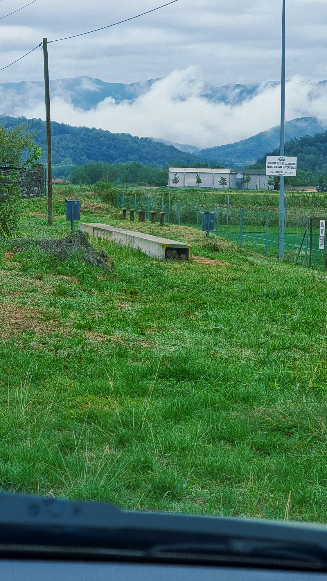 The image depicts a park-like setting in La Canya, Spain, with rolling, mist-covered hills in the background. In the foreground, a grassy area is sparsely populated with objects. A concrete structure, possibly a bench or low wall, runs horizontally across the mid-ground. To the left of this structure, a large rock or natural formation is visible, and to its left, a gray trash can stands. Further to the right, behind the concrete structure, a series of wooden benches are arranged. Another gray trash can is situated to the right of the wooden benches. A tall pole, likely a light post, stands near the right edge of the image, and attached to it is a white sign with text in Spanish. Beyond the grassy area, a fence encloses what appears to be a sports field with green turf. In the far background, several low-rise buildings are visible, nestled among trees. The sky is overcast, contributing to the subdued lighting.