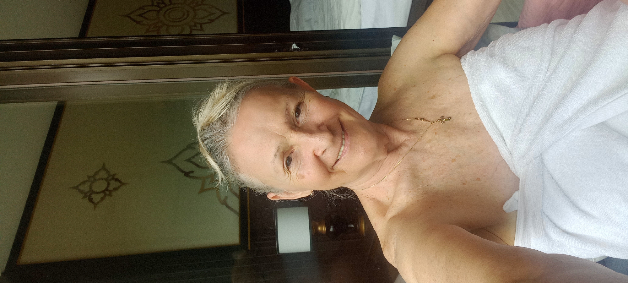 A person is shown from the shoulders up, wrapped in a white towel, smiling at the camera. They have white hair pulled back, a gold necklace with a cross pendant, and freckles on their chest and shoulders. Behind them is a window or glass door with a dark brown frame. Decorative floral designs are visible on the wall through the glass. The location is Ban Ao Nang, Thailand, suggested by the Thai-style floral design on the wall.