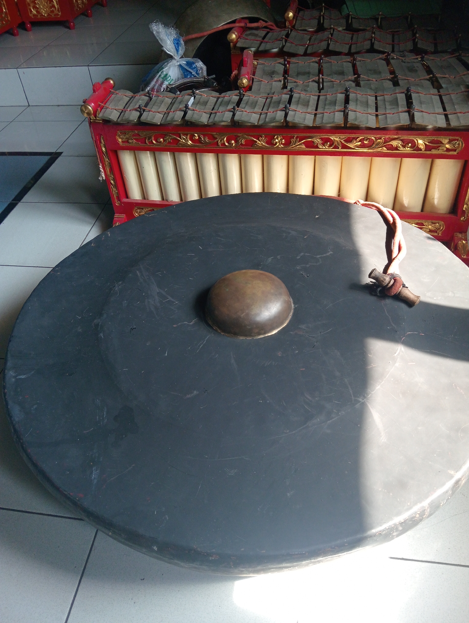 In Demak, Indonesia, an outdoor scene features a traditional Javanese gamelan instrument, specifically a metallophone with bronze bars, resting on a red and gold carved wooden frame. Next to it, a large, dark gong lies flat on the floor, with a hammer attached to a rope lying on its surface. The instruments are situated on a tiled floor, with a portion of a white wall visible in the background. Sunlight casts shadows across the gong.