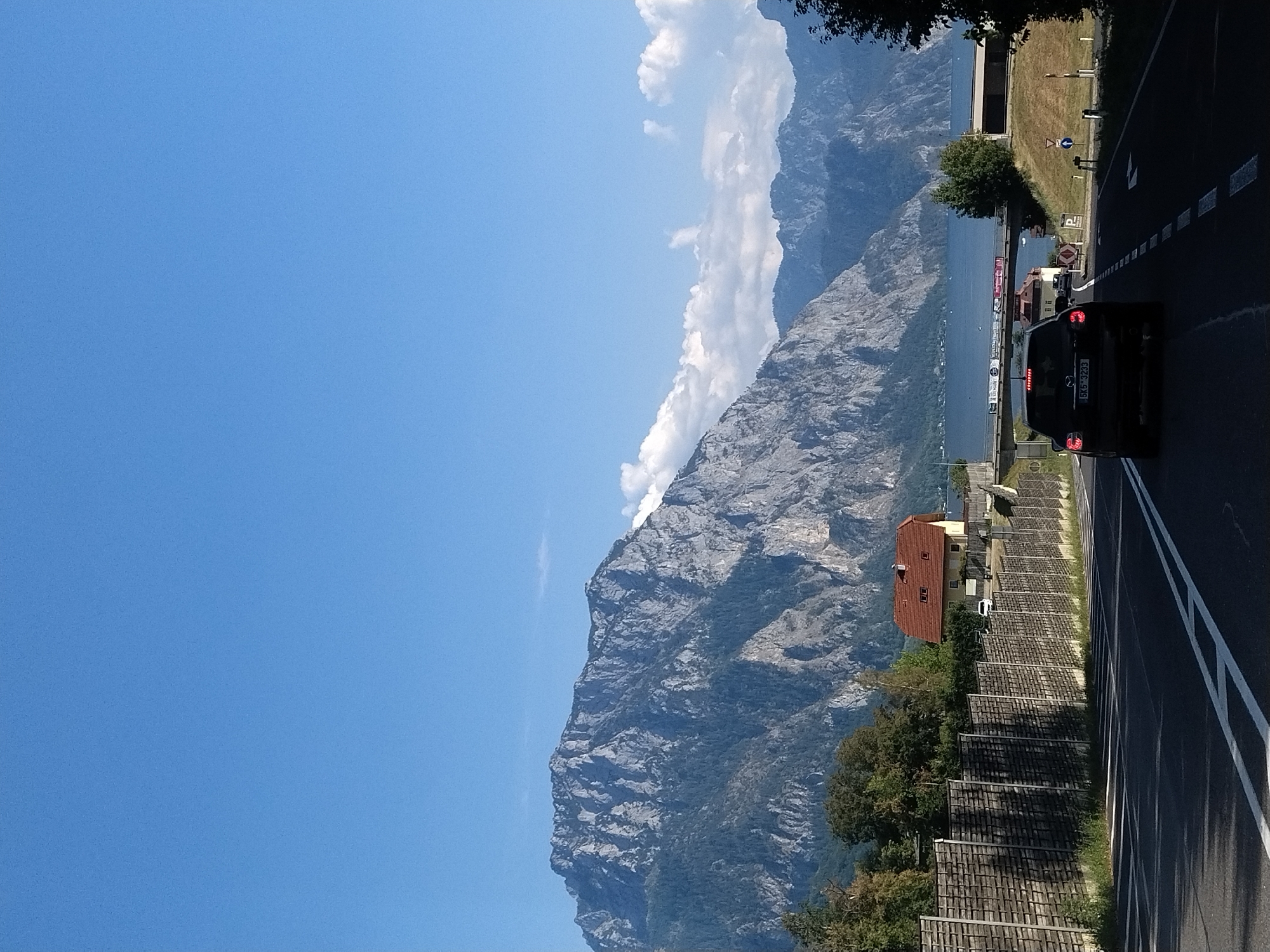 The image captures a scenic view of the Traunsee lake and the surrounding mountains, specifically the Traunstein, as seen from the road leading into Gmunden, Austria. In the foreground, a black car with license plate 5K5 3233 is driving on a paved road with white lane markings. A wooden fence runs along the right side of the road. A red-roofed house with light yellow walls is visible behind the fence. Further in the distance, the Traunsee lake is visible, with boats dotting its surface. The imposing Traunstein mountain dominates the background, its rocky slopes covered in dark green trees. The sky is a clear blue with a few scattered white clouds. The overall impression is a picturesque landscape scene, likely captured on a sunny day.