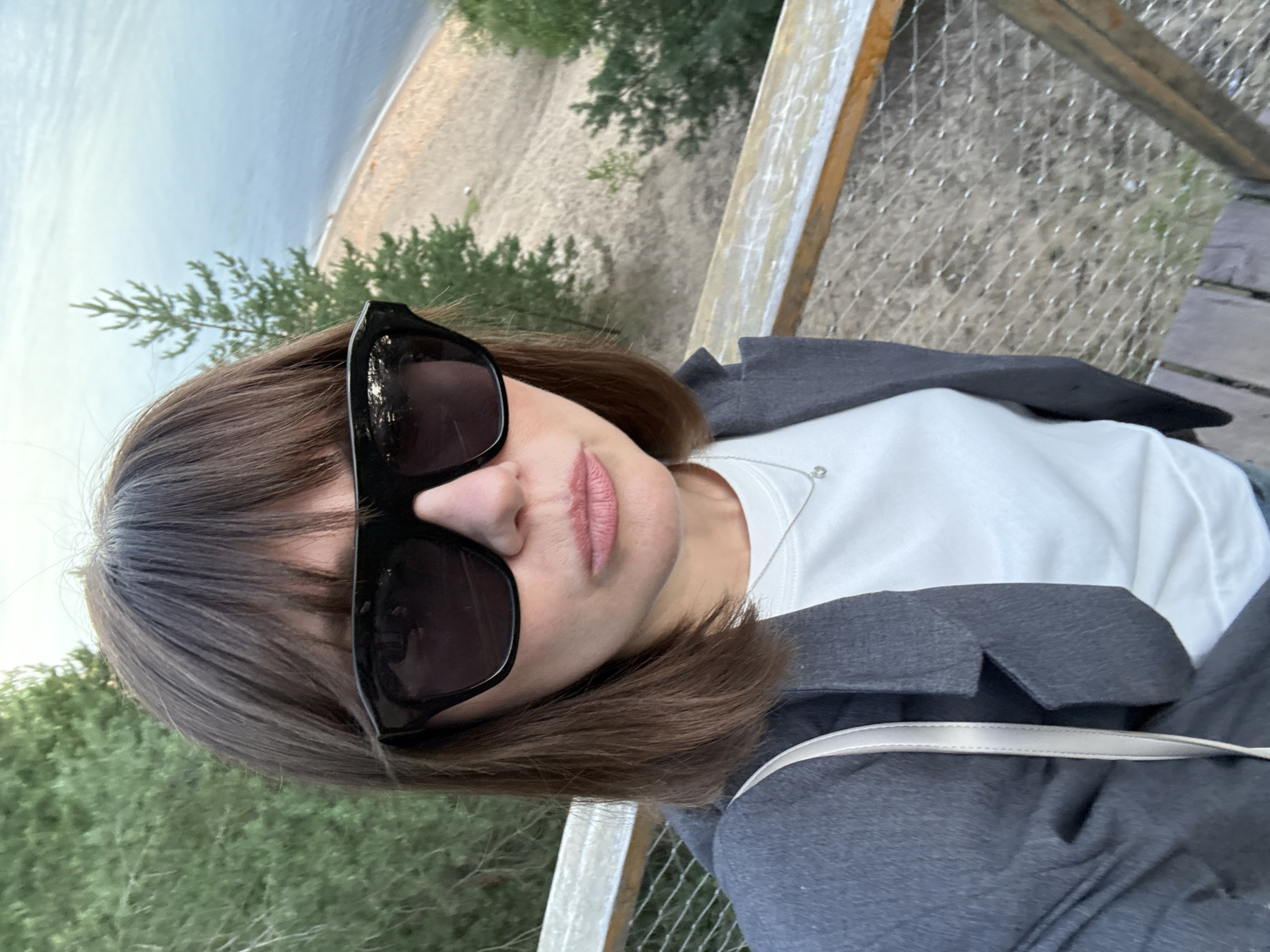 A woman with shoulder-length brown hair and bangs is taking a selfie outdoors. She is wearing large black sunglasses, a grey blazer, and a white t-shirt with a delicate silver necklace. Her lips are slightly parted. She is standing on a wooden platform with a metal mesh railing, overlooking a sandy beach and a body of water. Pine trees are visible in the background. The location is the observation deck in the city of Bor, Russia, known for its pine forests and proximity to the Volga River.