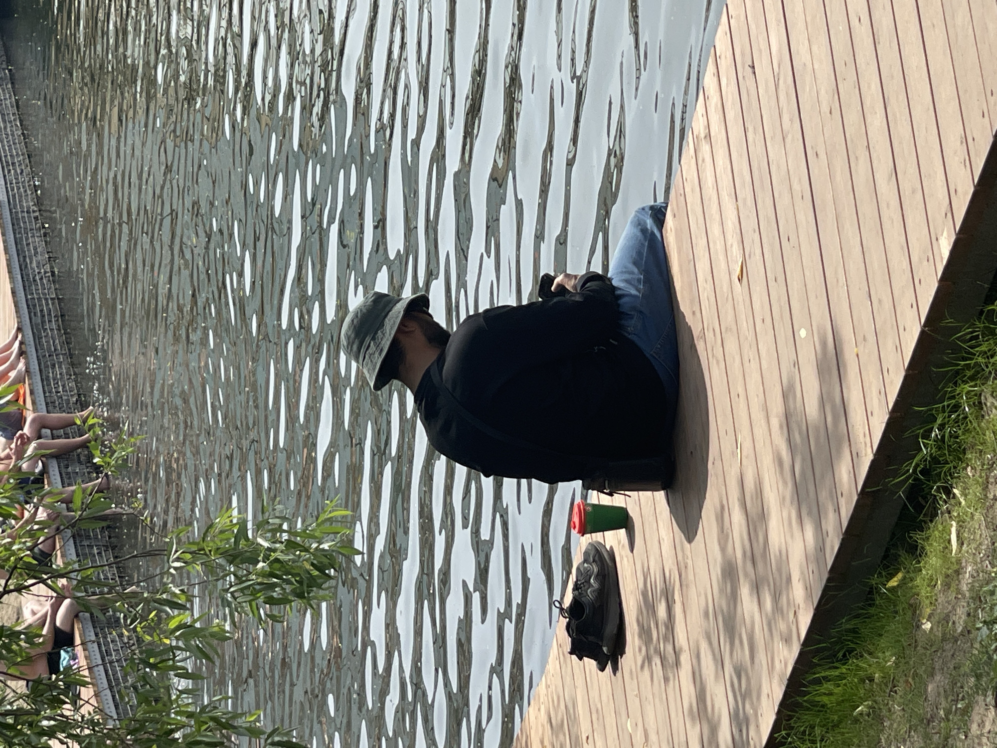 This eye-level outdoor photograph, taken in Moscow, Russia, captures a man seated on a wooden dock beside a body of water under bright daylight. The man is viewed from the back, wearing a dark green bucket hat, a long-sleeved black shirt, and blue jeans. He has a dark beard and is holding a dark-colored smartphone with both hands, looking down at the device. A black bag is positioned next to his right hip. To his left on the wooden planks of the dock are a pair of black athletic shoes and a green reusable cup with a red lid. The water, occupying the midground and background, exhibits ripples and reflections of sunlight. In the upper left background, several individuals are partially visible, seated on a similar wooden structure with their legs extended towards the water, partially obscured by hanging green tree branches. The wooden dock is composed of light brown horizontal planks. Green grass and bare earth are visible in the lower left foreground adjacent to the dock.
