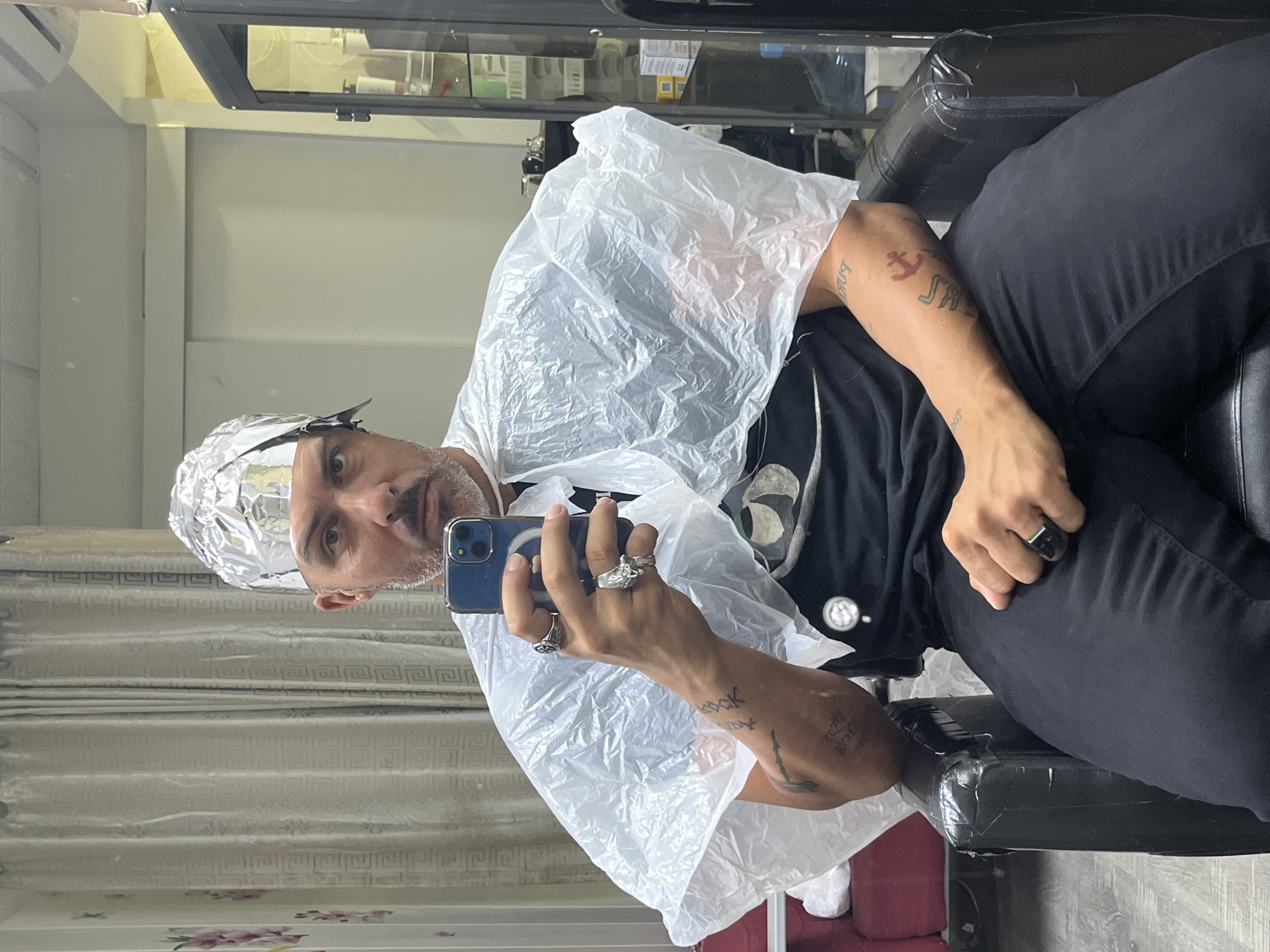 A man is taking a selfie in a hair salon. He is seated in a black chair, wearing black pants and a black t-shirt under a white plastic cape. His head is covered in foil. He is holding a blue smartphone in his left hand, which is adorned with rings. He has tattoos on both arms. The background shows light-colored curtains with a floral pattern and a mirrored wall, reflecting the salon's interior, including a display cabinet. The location is a hair salon that is likely in Barcelona, Spain.