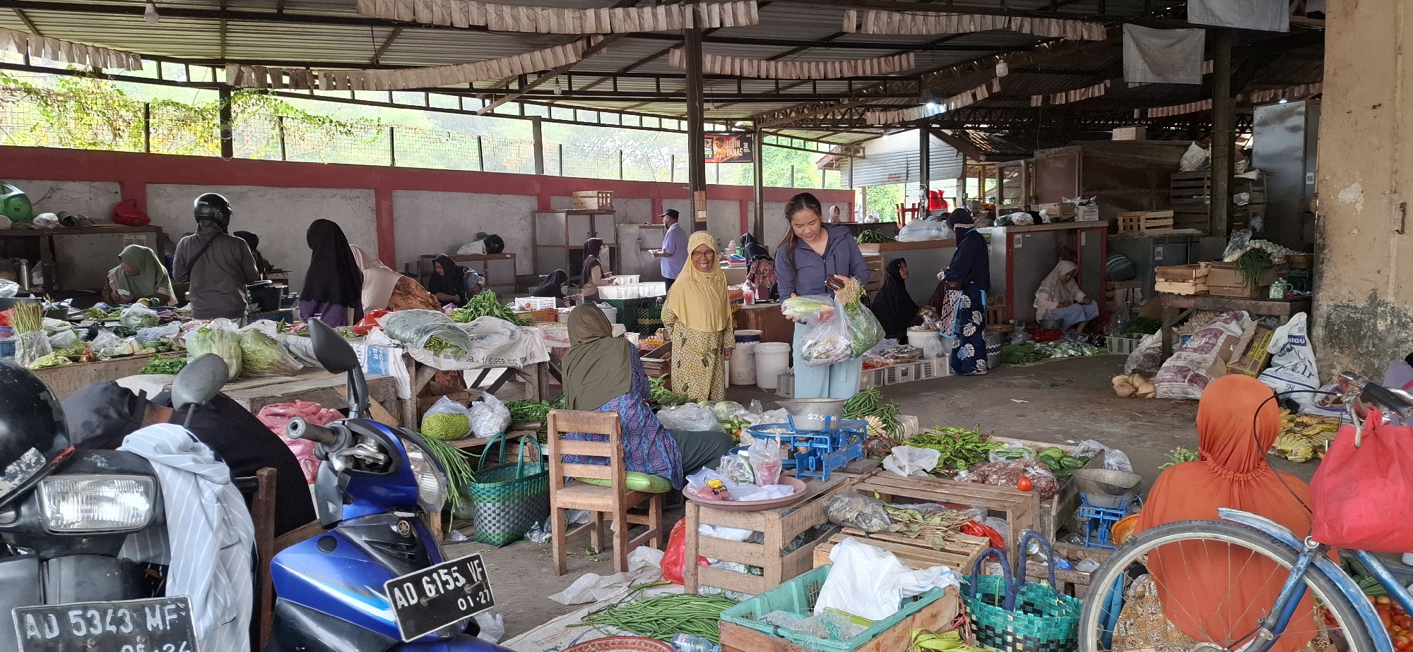 This is an outdoor market scene in Mranggen, Indonesia, likely a traditional market, with a covered stall area. In the foreground, two motorbikes are parked, one blue and one black, with their license plates visible: AD 6155 VF and AD 5343 MF. Various vegetables and produce are laid out on tables and on the ground, including leafy greens, tomatoes, bananas, coconuts, and what appear to be long beans.
Several people are present, mostly women, dressed in traditional Islamic attire. One woman in a yellow hijab is smiling and looking towards the camera. Another woman in a blue jacket is holding a bag of groceries. Other women are seated behind their stalls, attending to customers or arranging produce.
The market is organized with wooden stalls and tables, some covered with tarps. Crate boxes are used for displaying goods, and baskets are visible. The background shows a wall and some vegetation, suggesting an open-air setting within a built structure. The overall atmosphere is busy and typical of a local Indonesian market.
