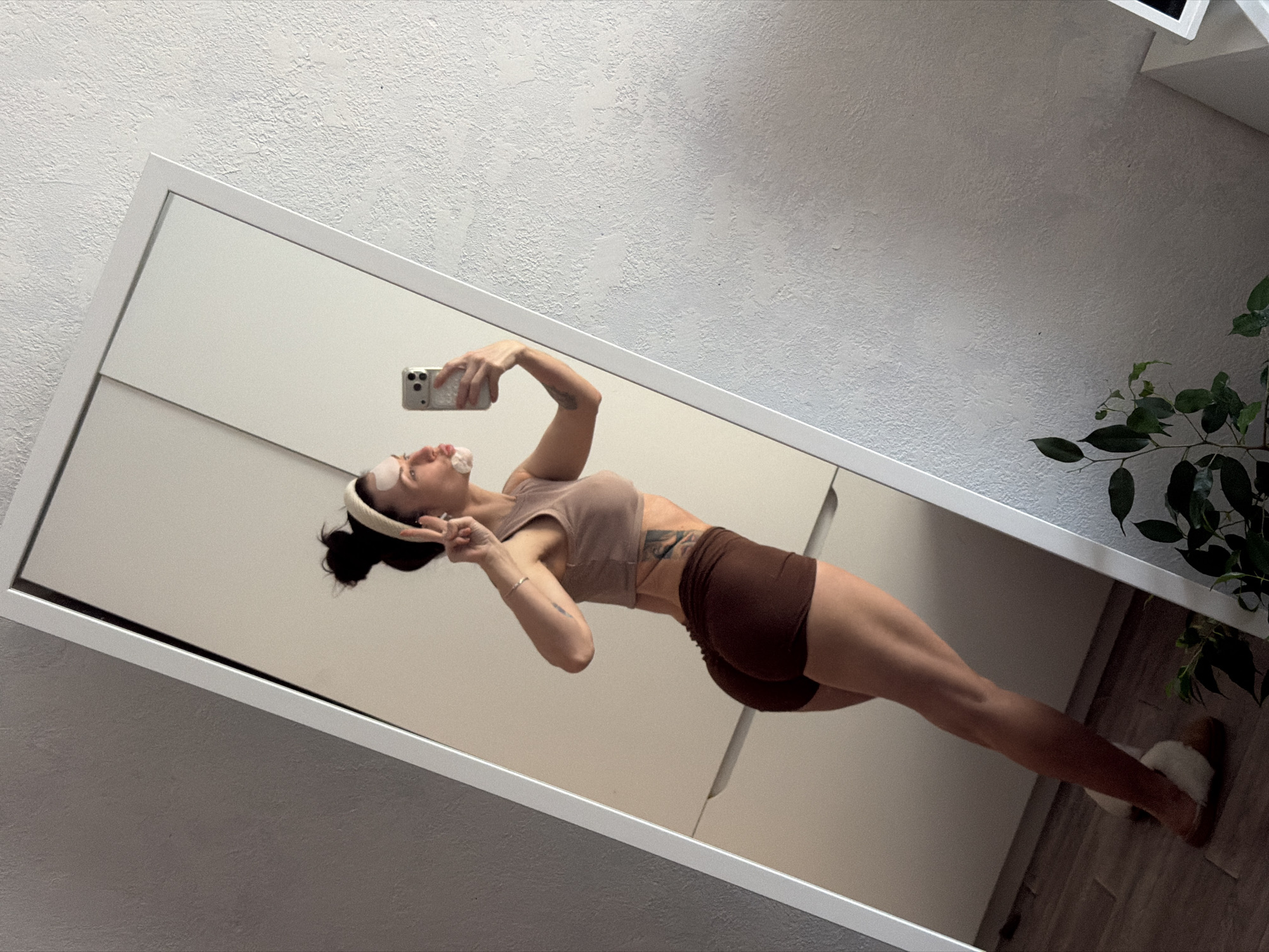 A full-body mirror selfie captures a woman posing in what appears to be a room in Perm, Russia. The image is taken at an angle, with a large, white-framed mirror dominating the scene. The woman, likely in her 20s or 30s, is reflected in the mirror. She has dark hair tied in a bun, wears a cream-colored headband, and has white patches under her eyes, suggesting a self-care routine. She is dressed in a light brown athletic crop top and dark brown athletic shorts, revealing a visible tattoo on her side torso. On her feet are fluffy white slippers.
She holds a silver smartphone with three camera lenses in her right hand, aiming it towards the mirror to capture the photo. Her left hand is raised, making a peace sign, and her face is turned slightly upwards with a playful pout or kissy expression. The mood conveyed is confident and relaxed.
The background outside the mirror's frame features a textured white wall (possibly stucco or rough plaster) on the upper left and a dark green leafy houseplant on the upper right. The reflection within the mirror shows a plain, light-colored wall and a wooden floor beneath her feet. The lighting suggests an indoor setting during the daytime or a well-lit interior. There is no visible text in the image.