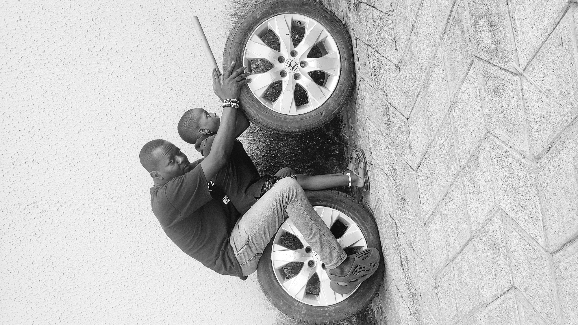A monochrome, vertically oriented image, captures a scene of an adult male interacting with a young boy in what appears to be Narayi, Nigeria. The adult, positioned on the left, is seated on a car tire, his legs in jeans resting over it, and his feet in patterned clogs touching the ground. He wears a dark short-sleeved top and has a focused expression as he holds the young boy in front of him.
The boy, wearing a dark top, shorts, and sandals with beaded ankle bracelets, is lifted by the adult. His hands are placed on another car tire that stands upright to his right. The boy also grasps a long, thin stick that extends upwards from the tire. He wears beaded bracelets on his wrists. The tire the boy interacts with features a silver rim with a visible Honda logo at its center.
The setting consists of a textured, stippled white wall on the left, forming the background behind the adult and partially behind the boy. To the right, a paved surface with a distinct herringbone pattern runs vertically, serving as a backdrop for the tires and part of the activity. The bright lighting suggests it is daytime. The interaction appears playful and supportive, with the adult assisting the boy.