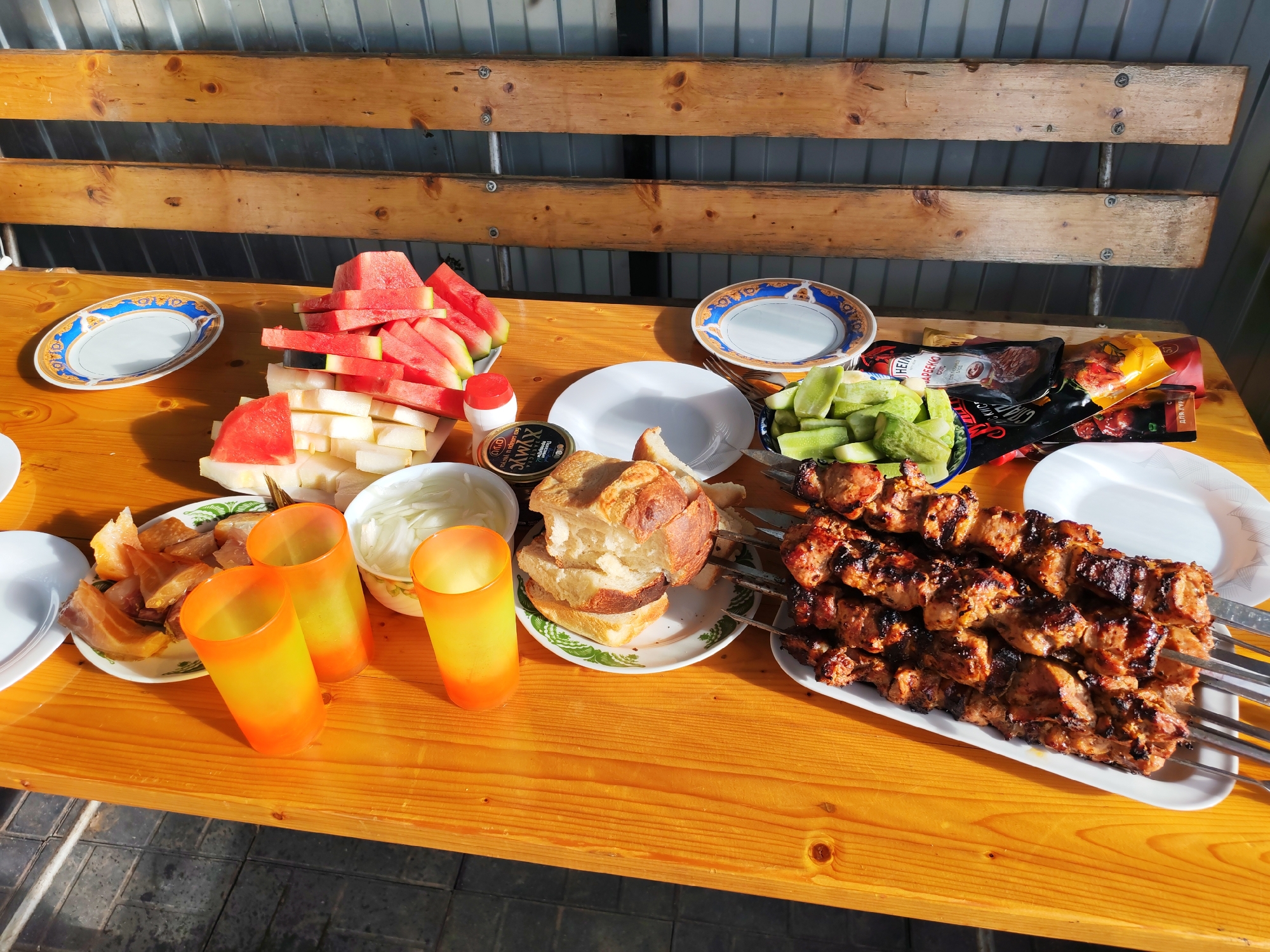 In Pervomaysky, Russia, a wooden table is laden with a spread of food. In the foreground, three vibrant orange and yellow gradient glasses are placed near a plate of smoked fish. Beyond them, a bowl of sliced onions, a plate of bread, and a bowl of chopped cucumbers are visible. The centerpiece of the table features several skewers of grilled meat, glistening and browned from the grill. To the left, a pile of sliced watermelon and melon sits on a platter, next to decorative plates. The backdrop consists of a corrugated metal fence and a wooden slatted shelf. The scene suggests a casual outdoor meal or gathering.