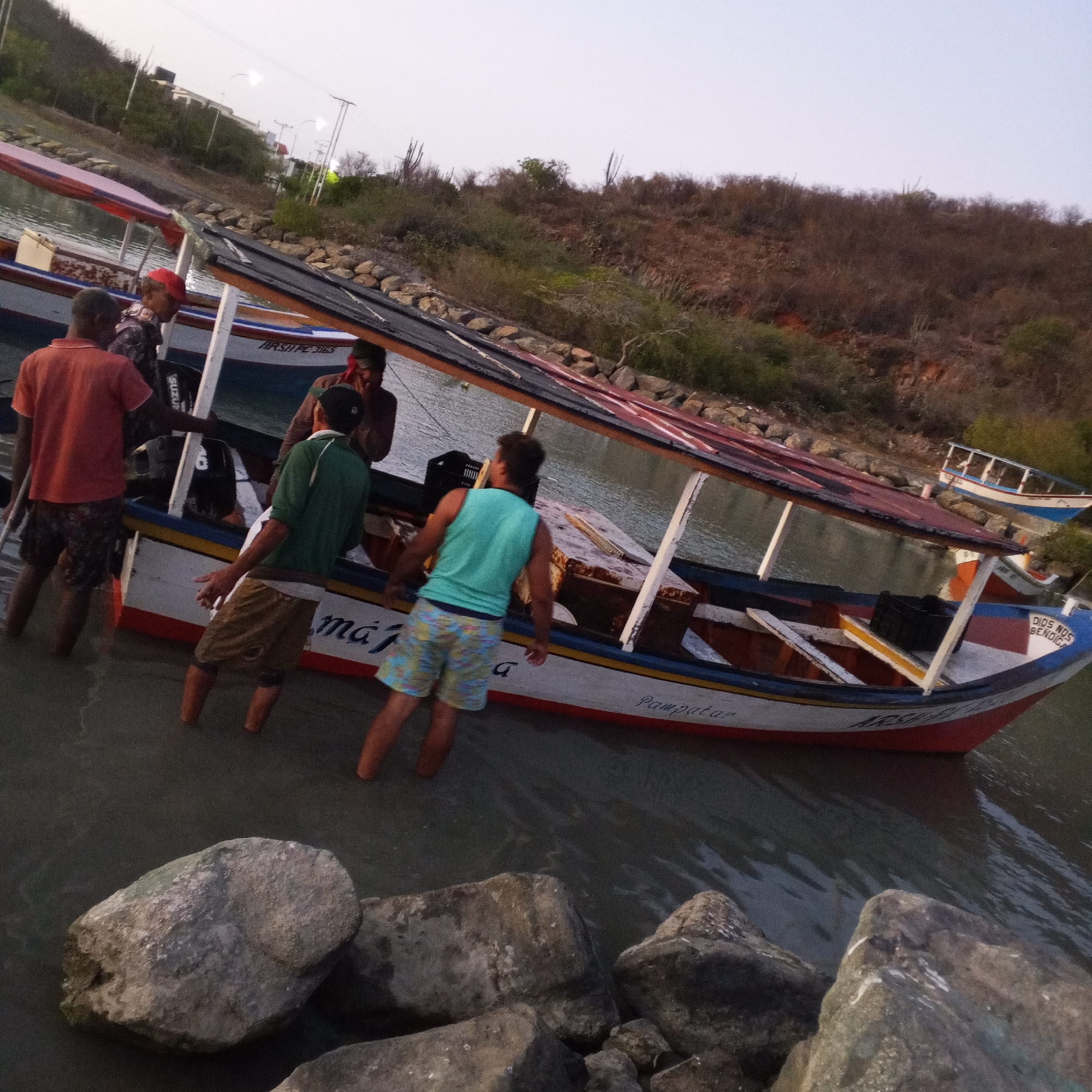 The image shows a group of men standing in shallow water near a boat, possibly in the coastal city of Pedro González, Venezuela. The boat, which appears to be a small fishing vessel or water taxi, has a roof and is decorated with the words "Pampatar" and "Dios Nos Bendiga". The men are casually dressed and seem to be interacting with the boat. The surrounding environment includes a hillside with vegetation and some rocks in the foreground.