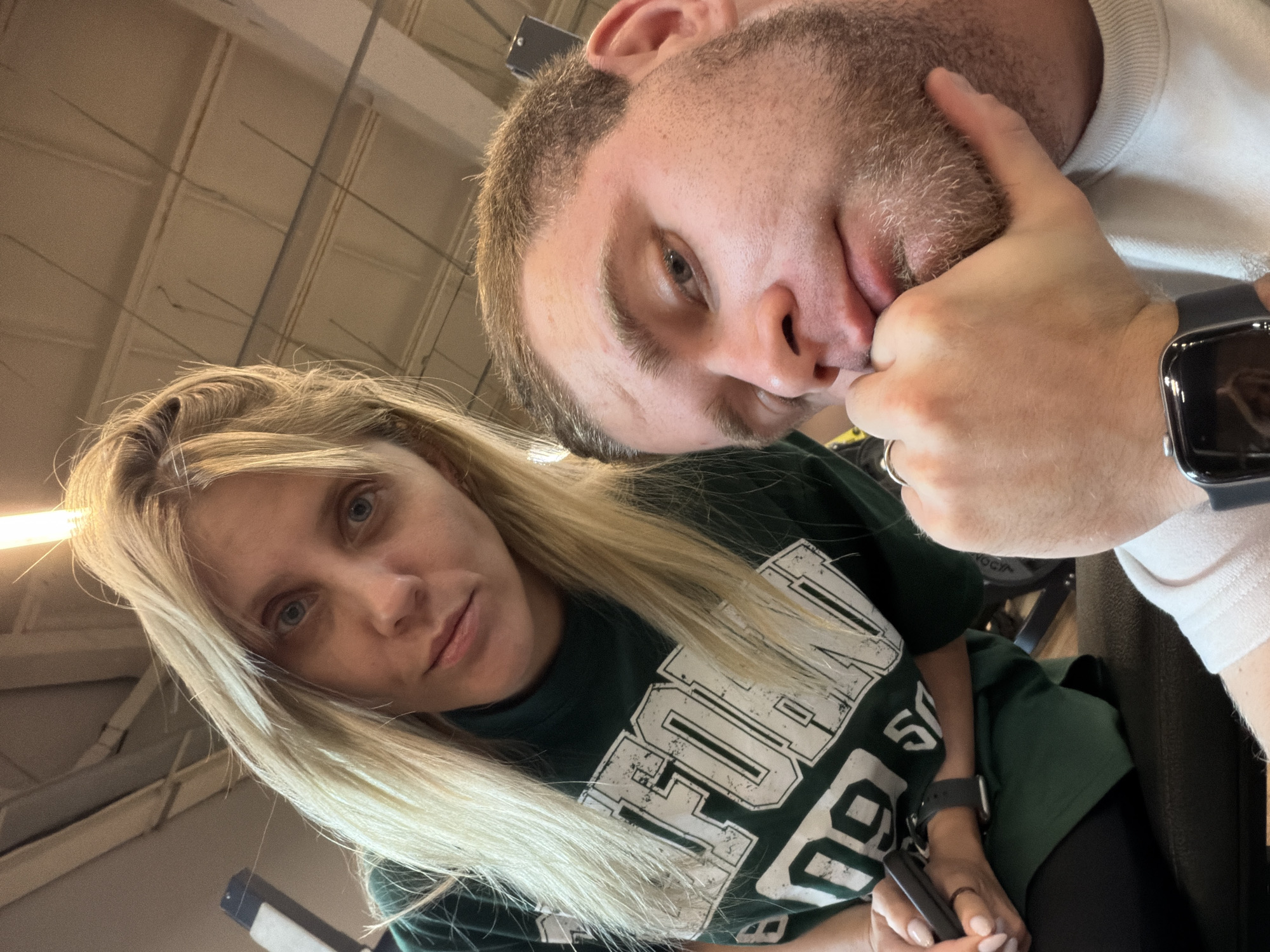 A man and a woman are posing for a selfie in what appears to be a gym. The woman, with blonde hair, is wearing a green t-shirt with white lettering. The man, with short brown hair and a beard, is resting his chin on his hand. The background shows gym equipment and a ceiling with lighting. The scene is captured from a low angle, looking up at the subjects. The image does not contain enough information to determine the location in Chaikovsky, Russia.