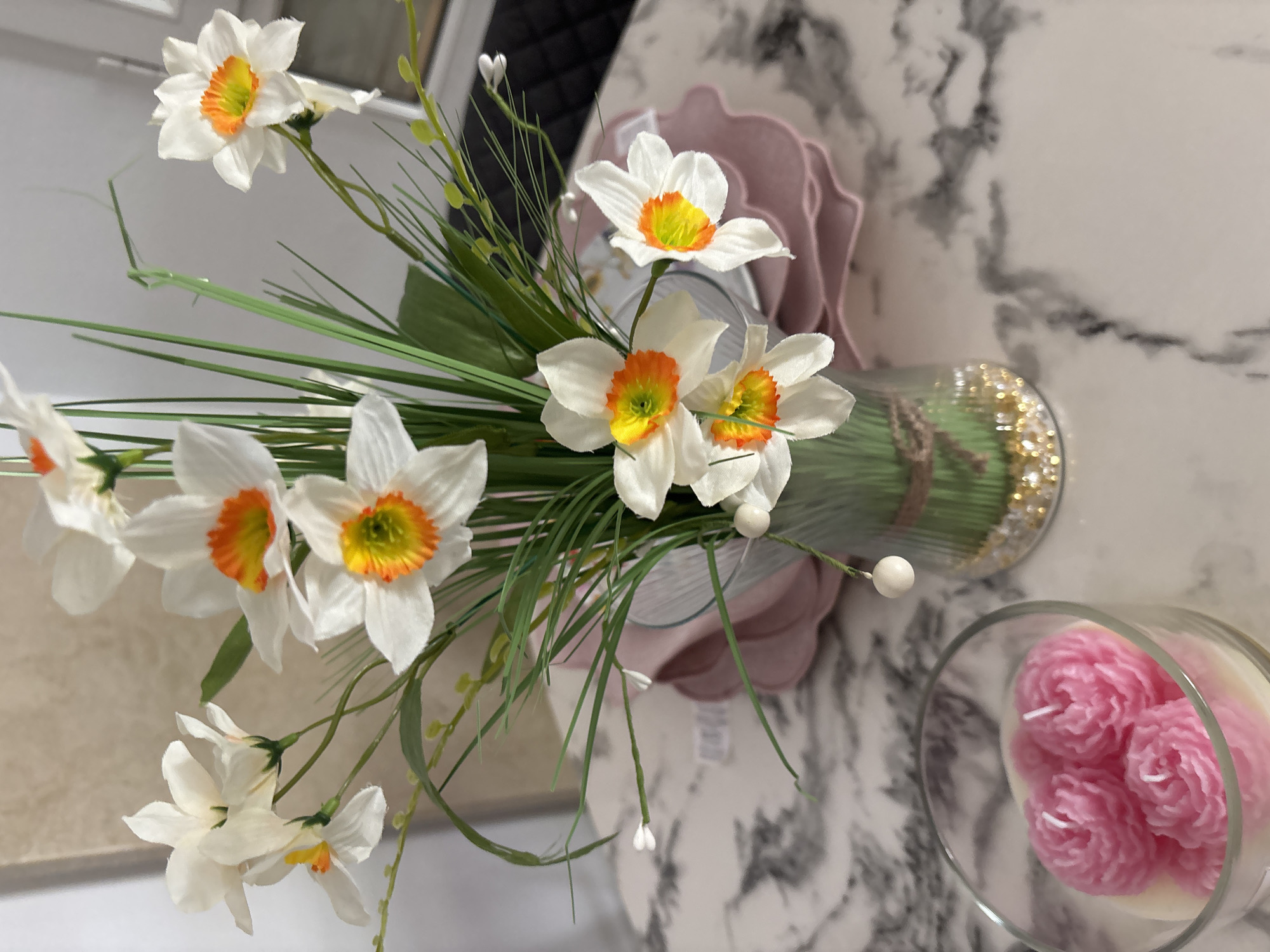 An arrangement of artificial white daffodils with orange centers is displayed in a clear glass vase on a marble-patterned table. The vase contains green artificial grass and gold and white decorative beads at the bottom. To the left of the vase, a clear glass container holds three pink, rose-shaped candles. In the background, stacked pink plates are visible. The image does not provide information about the city of Orihuela, Spain.