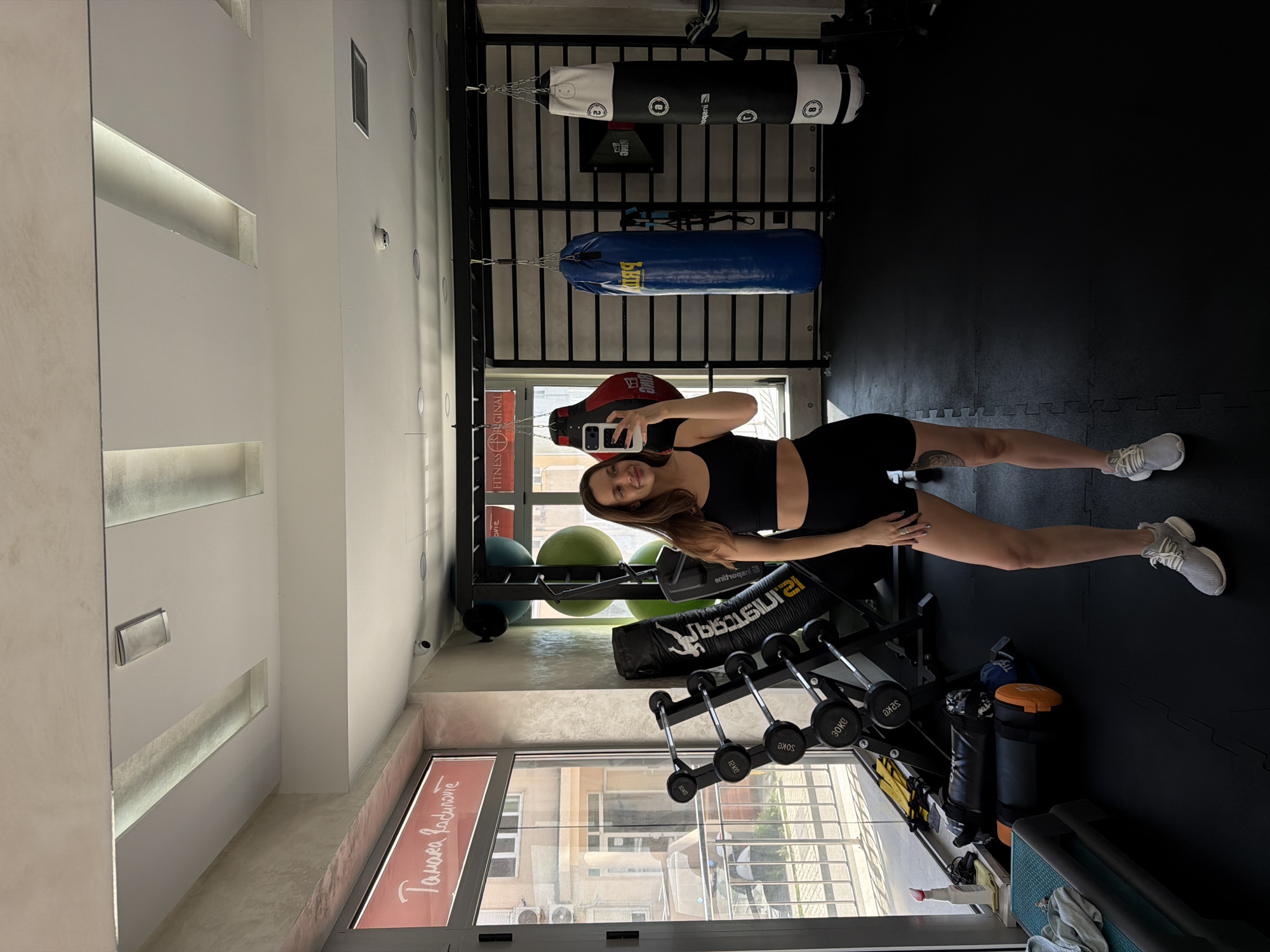 A woman stands in a gym, holding a smartphone, likely taking a selfie. She is wearing a black athletic top and shorts, and white sneakers. The gym appears to be well-equipped with various workout gear, including punching bags suspended from a frame, a rack of dumbbells, and exercise balls. Through a window, buildings can be seen. There is a sign on the window that reads "Тамara Радоnovіć". A search reveals that this is likely the "Tamara Radinović Fitness Original" studio in Budva, Montenegro. The gym has white walls and black flooring.