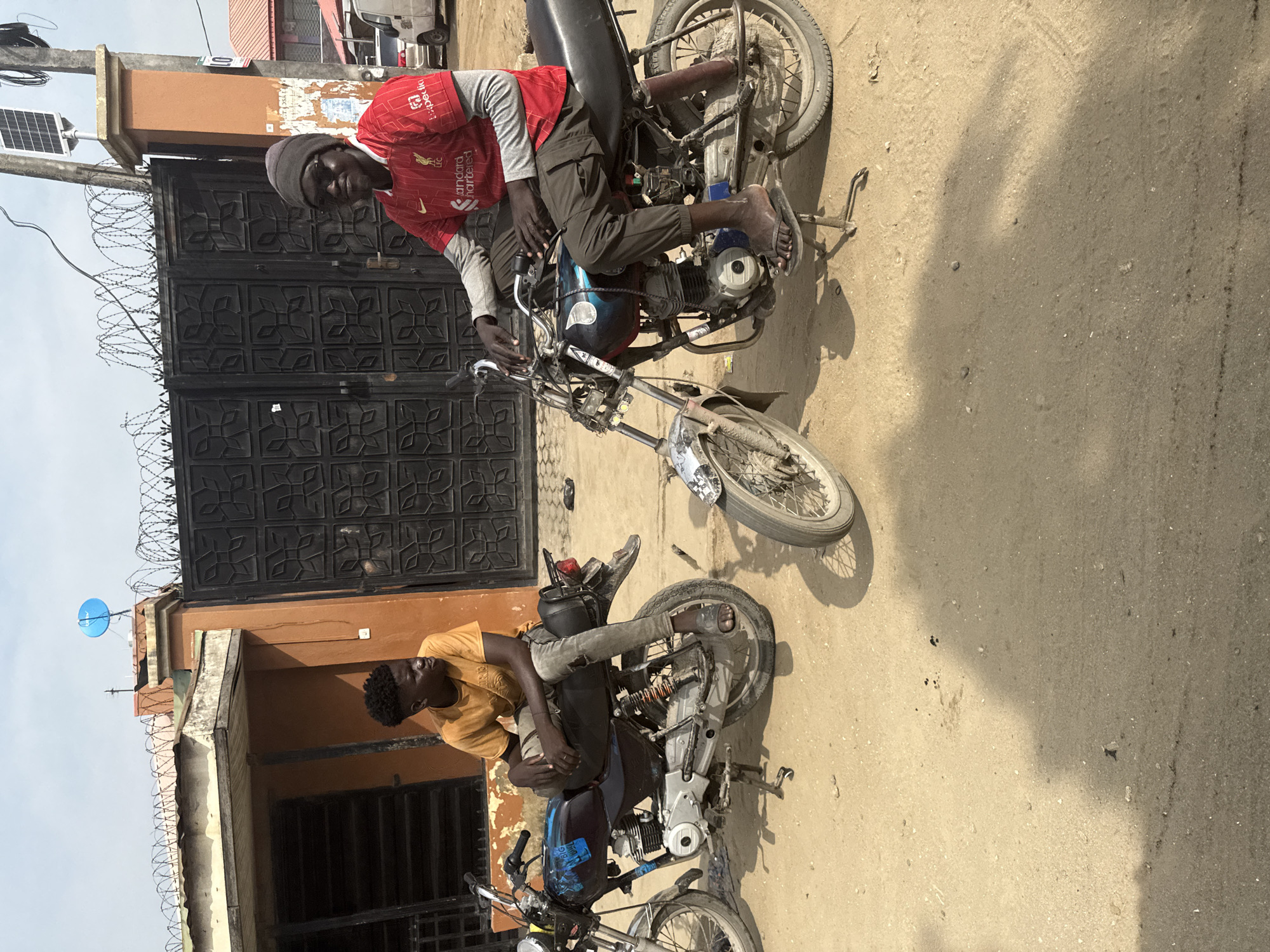 The image, captured in Ado, Nigeria, depicts an outdoor scene with two young men and two motorcycles on a dusty, unpaved surface under bright daylight. The image is oriented vertically, rotated 90 degrees clockwise from a natural upright perspective.
In the mid-ground, a young man sits on a black motorcycle. He wears a red long-sleeved football jersey featuring "Standard Chartered," "Expedia," "AXA," and the "Liverpool FC" crest, along with the text "NB". He also has on a grey beanie, dark cargo-style pants, and flip-flops. He looks directly towards the camera with a neutral expression. The motorcycle appears functional but visibly worn.
Further back, another young man leans against a building next to a second black motorcycle. He is dressed in a yellow short-sleeved t-shirt, grey pants, and sandals. He looks away from the camera, towards the left of the frame (when viewed upright). This motorcycle also shows signs of use. Both men are dark-skinned.
The background features a dark, patterned metal gate topped with barbed wire, behind the first man. Adjacent to the gate, a pole supports a small solar panel. A brown-painted building with barred windows and more barbed wire along its roofline is visible behind the second man. A bright blue satellite dish is mounted on a building in the far background. The ground is a light-colored, dusty or sandy terrain, with strong shadows indicating a sunny day, likely mid-morning or afternoon. Faint, mostly illegible posters or markings are on the walls, with a clear number "19" visible on a reddish pole near the first man. The overall scene suggests a casual, everyday setting in an urban or semi-urban environment.