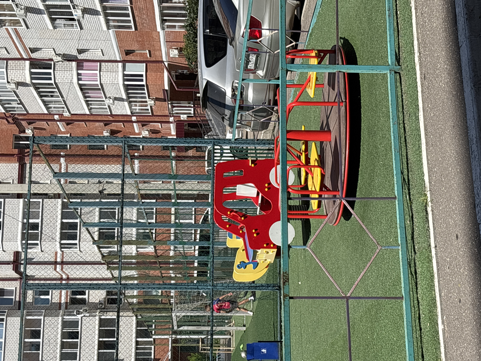 The image shows a children's playground with a red and yellow car-shaped ride in the foreground, and a red and yellow carousel to its right. The playground is on a green artificial turf surface, enclosed by a green metal fence. In the background, a young person wearing a red and blue shirt with the number 19 is visible, walking across a sports field with a soccer goal. Beyond the sports field, a multi-story brick building is visible, with multiple windows and air conditioning units. Two silver cars are parked behind the fence. A blue trash can is located to the left of the playground. The scene appears to be outdoors on a sunny day. This is a residential area in Chigiri, Russia.