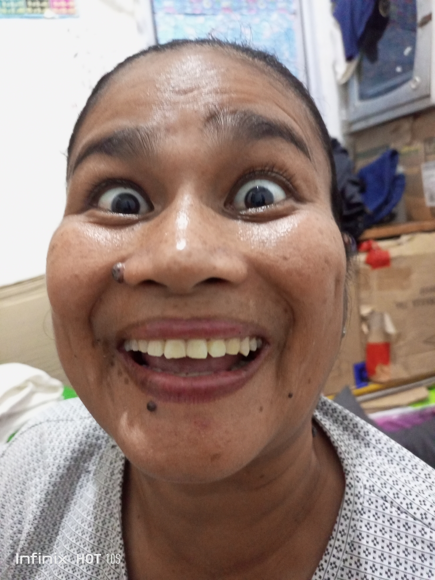 A close-up, frontal, eye-level, selfie photograph captures a woman making a humorous facial expression. Her eyes are wide and bulging, her eyebrows are raised, and her mouth is open, revealing her teeth in a wide smile. The woman has dark skin, short dark hair, and several moles on her face, including one on her nose. She is wearing a light-colored shirt with a pattern of dark, diamond-shaped symbols. The background is blurred and appears to be an indoor setting with shelves and various objects, including what looks like cardboard boxes and colorful items. The photograph was taken in Batam, Indonesia, as indicated by the "Infinix HOT 10S" watermark in the lower left corner, which is a model of smartphone often used in Indonesia.