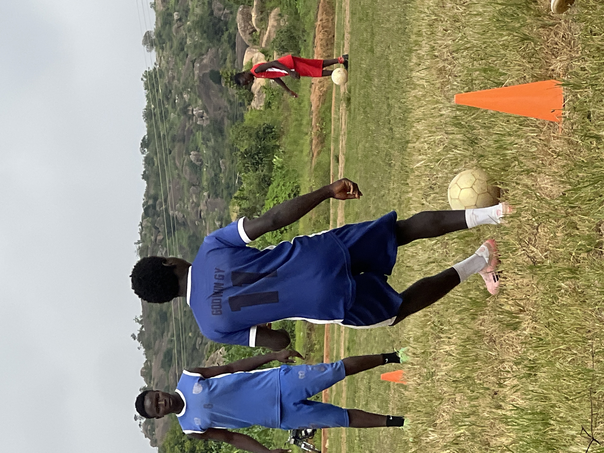 In Jos, Nigeria, three young men are engaged in a football training session on a grassy field. Two men in blue uniforms are visible, one in the foreground with his back to the camera, wearing a blue jersey with "GODWIN GY 11" printed on the back, and blue shorts. He is dribbling a light-colored football. Another man in a blue sleeveless jersey and blue shorts stands to his left, facing the camera. A third man in a red jersey and red shorts is further in the background on the right, also with a football at his feet. The environment features rocky hills and sparse vegetation in the background under an overcast sky. Orange cones are placed on the field, suggesting a training drill. A motorcycle is parked on the left side of the frame.