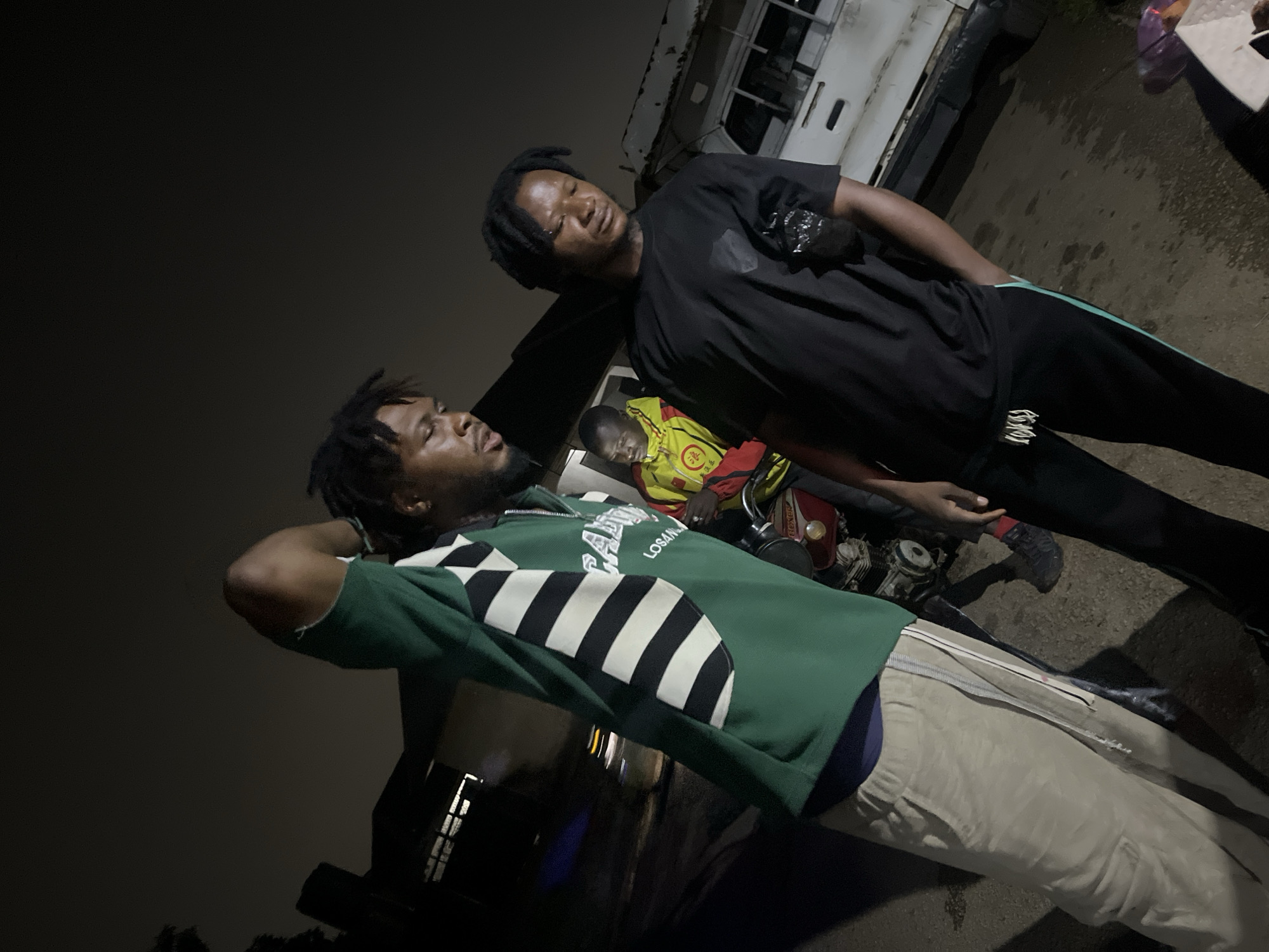 Three individuals are visible in this nighttime outdoor scene, likely in Jos, Nigeria. The primary focus is on two young men standing close together. The man on the left wears a green long-sleeved shirt with white and black striped sleeves and tan pants. He is looking down and to his right, with his left hand raised to his head. The man on the right, dressed in a black t-shirt and black track pants with a green stripe, is facing away from the camera, with his head bowed. A third person, wearing a yellow and red jacket, is partially visible in the background, seated on a red motorcycle. Behind them, a white truck with visible damage is parked. The ground appears to be paved, and there are some shadowy structures and a hint of vegetation in the background. The lighting suggests artificial illumination from an unseen source.