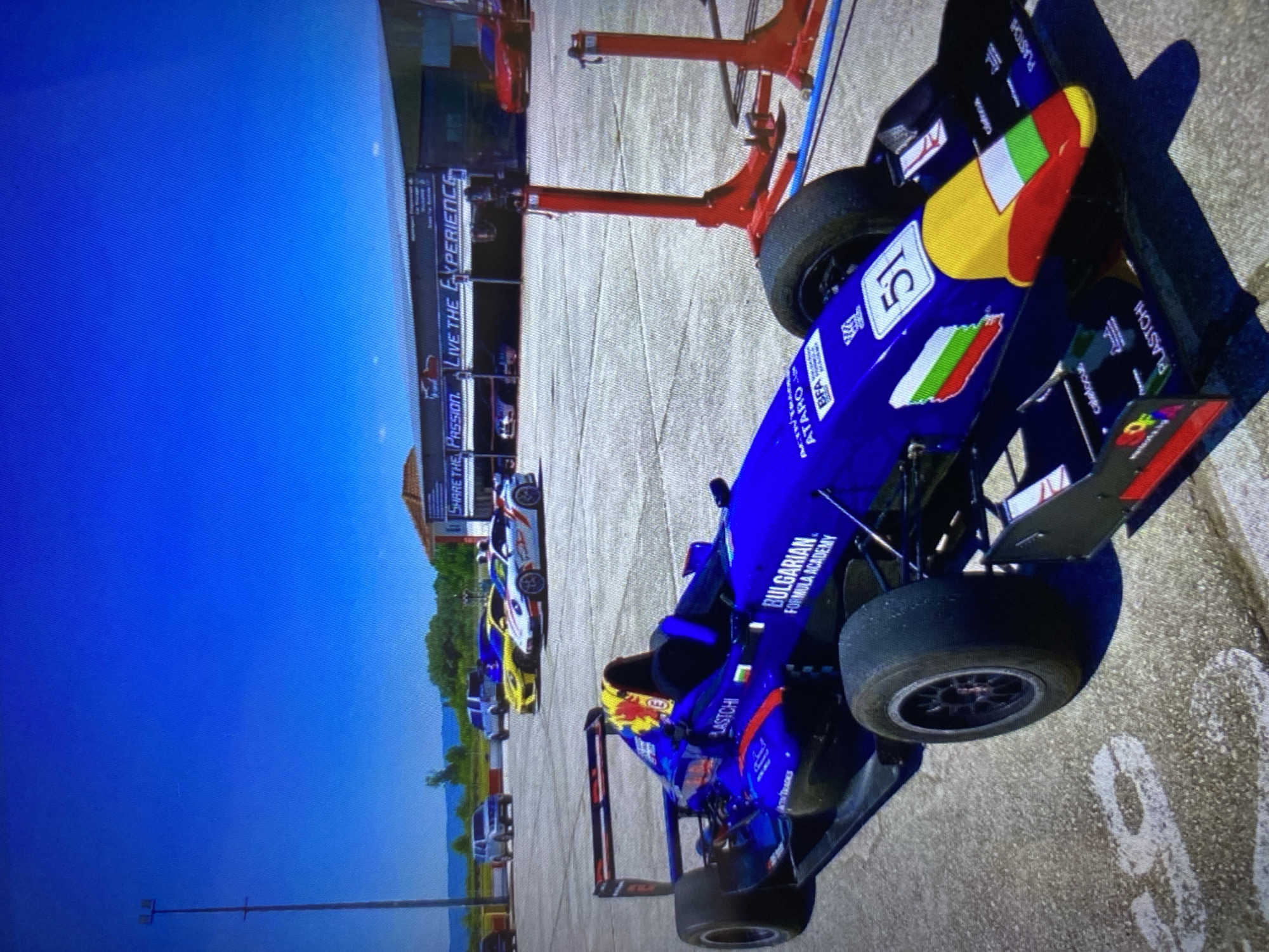 The image captures an open-wheel race car, likely a Formula car, positioned in what appears to be a pit area or paddock of a race track, under a clear, bright blue sky. The car, predominantly blue, features the number "51" prominently on its nose cone.
Visible text on the car includes "BULGARIAN FORMULA ACADEMY", "ATARO", "LASTCHI", "PLASTCHI", "BFA", "ACITRA", and "ATN TRADES", along with "calelocus". The car's design prominently incorporates Bulgarian national symbols, with the white, green, and red stripes of the Bulgarian flag on its nose and a map outline of Bulgaria with the flag on its side. This strongly indicates its affiliation with Bulgaria, specifically Sofia as per context.
In the background, other race cars in various colors (white, yellow, and red) are partially visible further down the track or pit lane. Red lifting equipment or stands are positioned behind the main blue car. A building in the background displays a banner with the text "SHARE THE PASSION. LIVE THE EXPERIENCE." The outdoor setting includes distant mountains or hills under the clear sky, indicating good weather conditions during the daytime. The ground is a light-colored concrete or asphalt surface. The scene suggests an active motorsport event or practice session taking place.