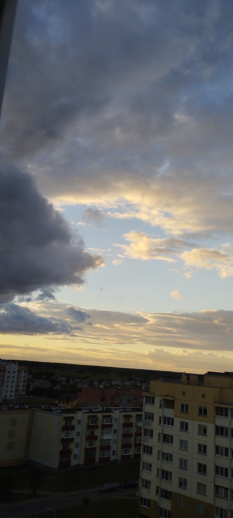 The image depicts a view of a city, likely Skidzieĺ, Belarus, from an elevated position, possibly a high-rise building. The sky dominates the upper portion of the frame, filled with dramatic, layered clouds. Dark, heavy clouds on the left transition into lighter, more diffused clouds with hints of yellow and orange, suggesting either dawn or dusk. The lower half of the image shows a panorama of urban development. Several multi-story apartment buildings are visible, characterized by their predominantly light yellow and white facades, with balconies and numerous windows. Some buildings have darker red accents. A distant tree line forms a dark silhouette against the brighter sky near the horizon. Below the apartment buildings, a road is visible with at least two dark-colored cars parked or driving. The overall scene conveys a sense of a residential area under a dynamic sky.