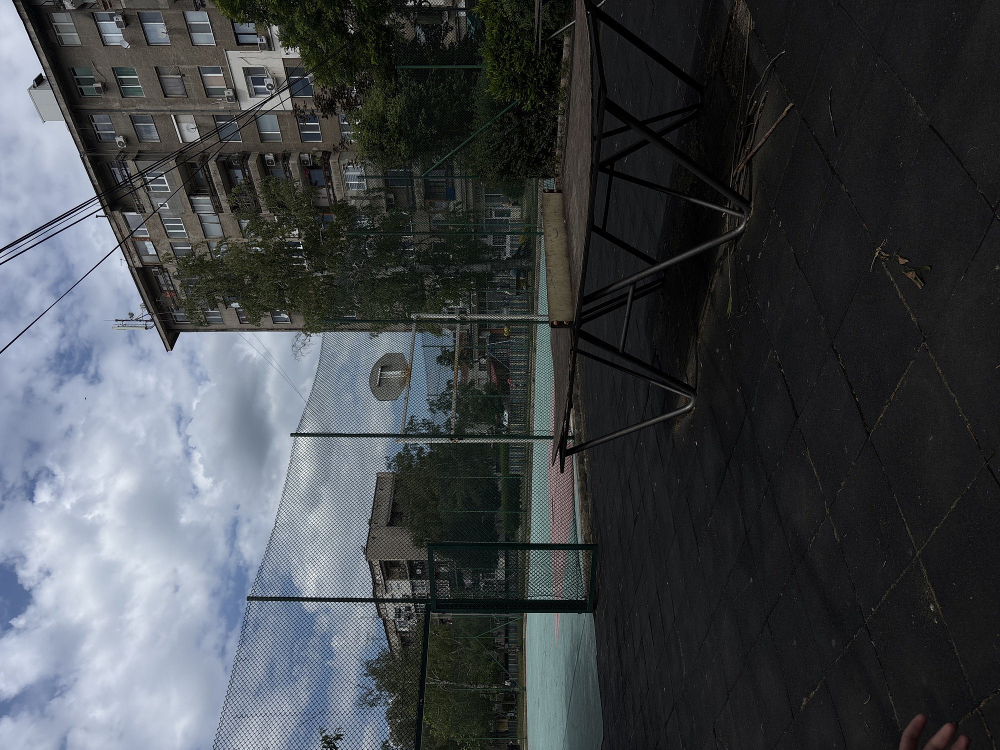 The image depicts an outdoor basketball court in Lom, Bulgaria, bordered by a chain-link fence. A multi-story residential building is visible in the background, partially obscured by trees. The court surface appears to be composed of dark, interlocking rubber tiles. A basketball hoop is visible through the fence. A metal structure, possibly a bench or another piece of equipment, is in the foreground. The sky is visible through the fence, with scattered clouds.