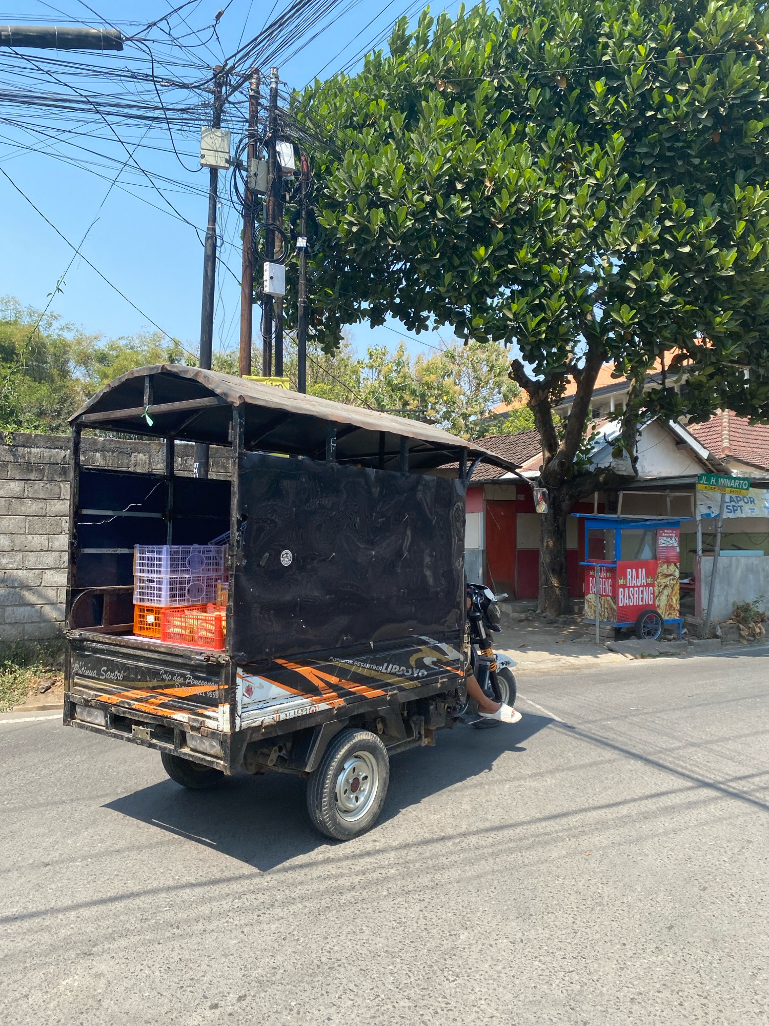 In Kediri City, Indonesia, a three-wheeled vehicle with a covered cargo bed is traveling on a paved road. The vehicle is loaded with white and orange plastic crates, some containing what appear to be fruits or vegetables. A person's legs are visible, indicating they are operating the vehicle. To the right of the road, there is a food stall with signage that reads "RAJA BASRENG". Further in the background, buildings and trees are present, and a complex network of overhead electrical wires spans the scene. The sky is clear and blue.