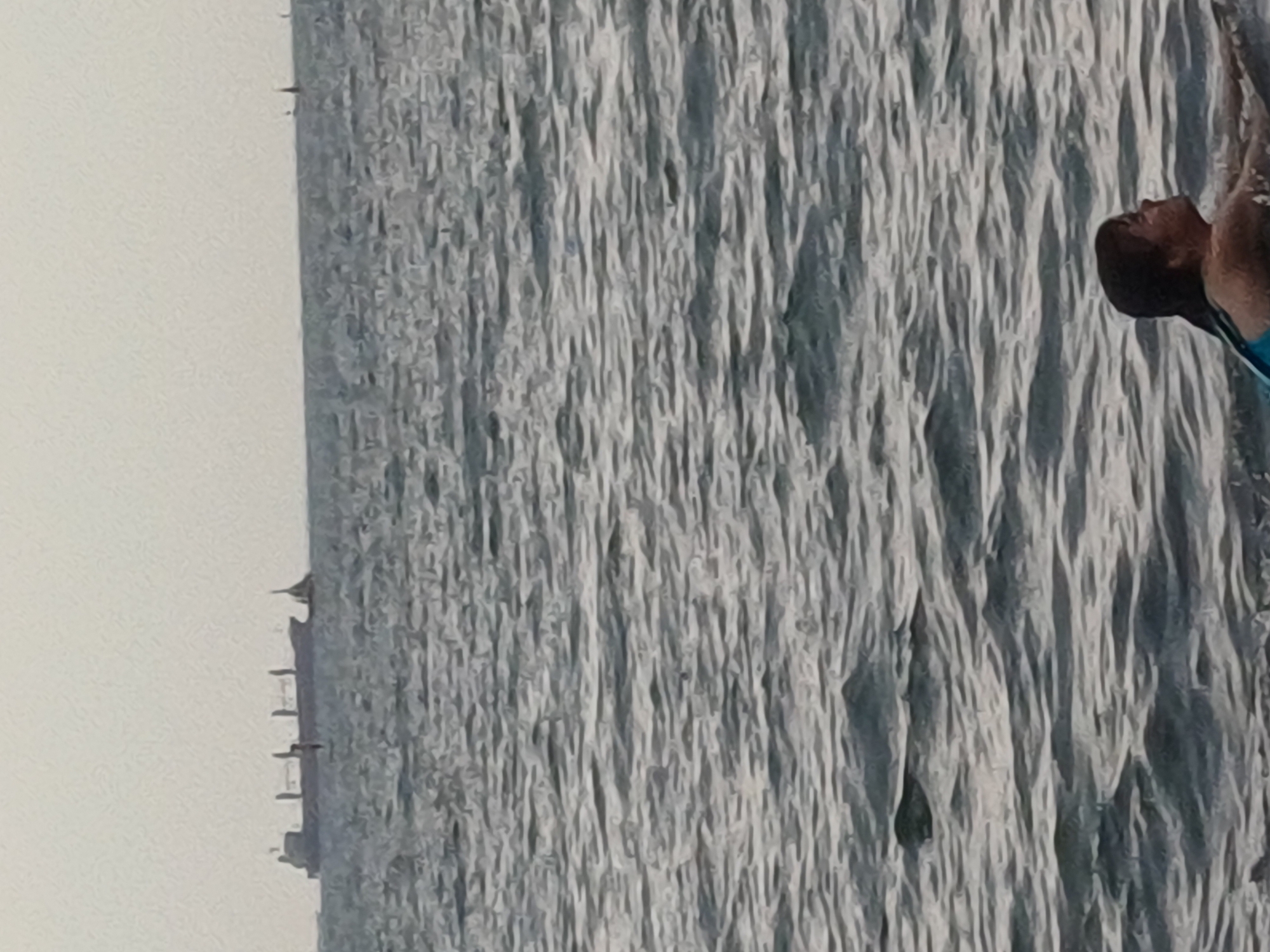 The image shows a person in the water with a blurred background of rippling water. The person appears to be male, with dark hair and is wearing a blue swimming garment. The person is looking upwards, with their head tilted back. The water has a textured appearance due to the waves and light reflecting on the surface. The overall tone of the image is muted, with shades of gray and blue dominating the color palette. The sky is not visible in the frame, but the lighting suggests it could be daytime. The scene appears to be set in a body of water, possibly the sea or a large lake, given the extent of the rippling water. In the far background, on the left side of the image, there is a faint outline of buildings, which might suggest an urban or coastal setting. No text is visible in the image. The overall mood is peaceful and serene, with the focus on the individual and their interaction with the water. The image is taken from a slightly elevated angle, looking down at the person in the water.