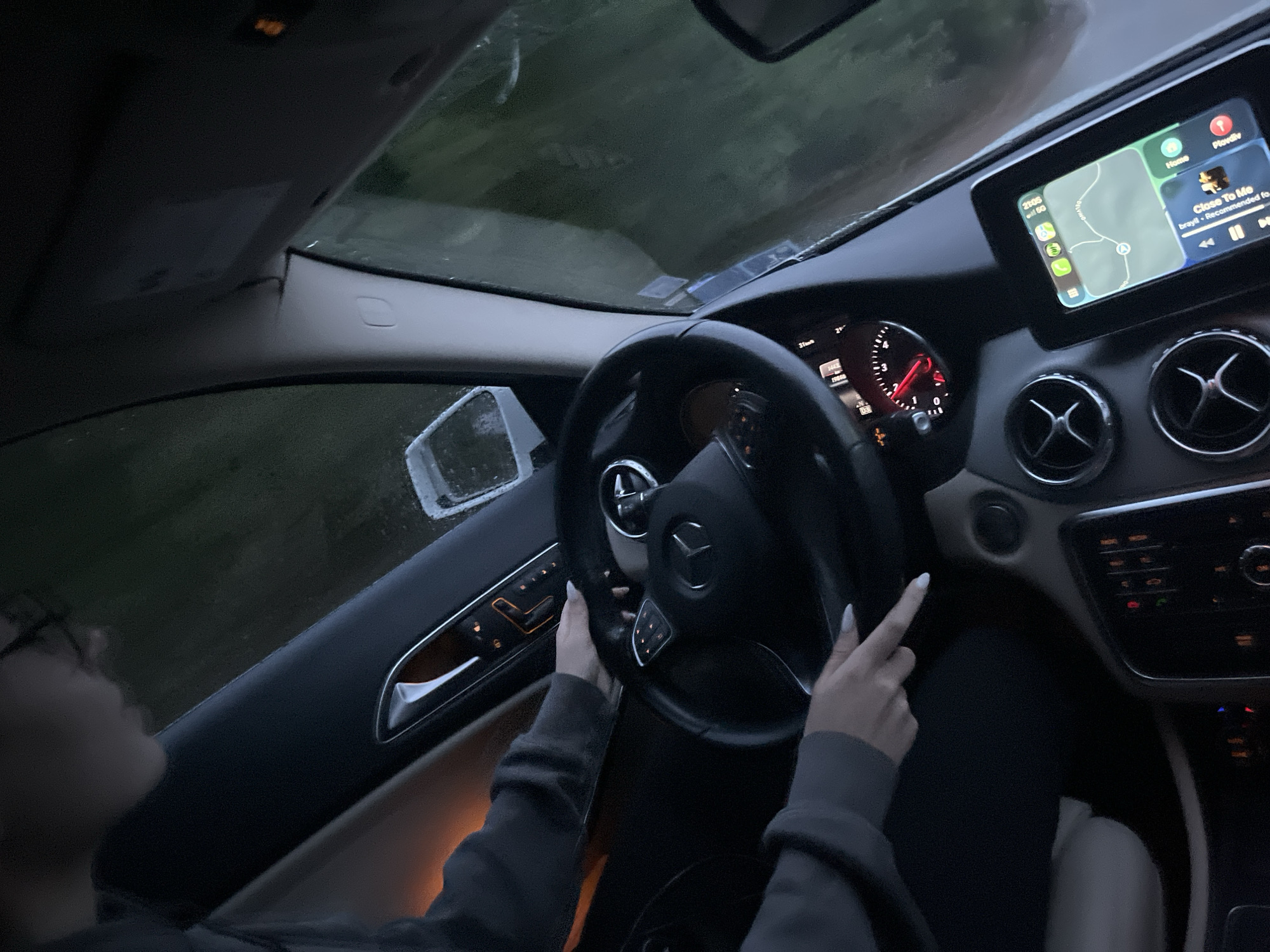 A person is driving a Mercedes-Benz vehicle. The car's infotainment system displays a map with a route highlighted. The speedometer needle is visible, showing a low speed. Rain or moisture is visible on the windshield and side mirror. The surrounding environment appears to be an outdoor area, possibly a road with vegetation alongside it. The GPS is showing an end point location in the city of Blagoevgrad, Bulgaria. The song "Close To Me" by brayll is playing on the infotainment screen.