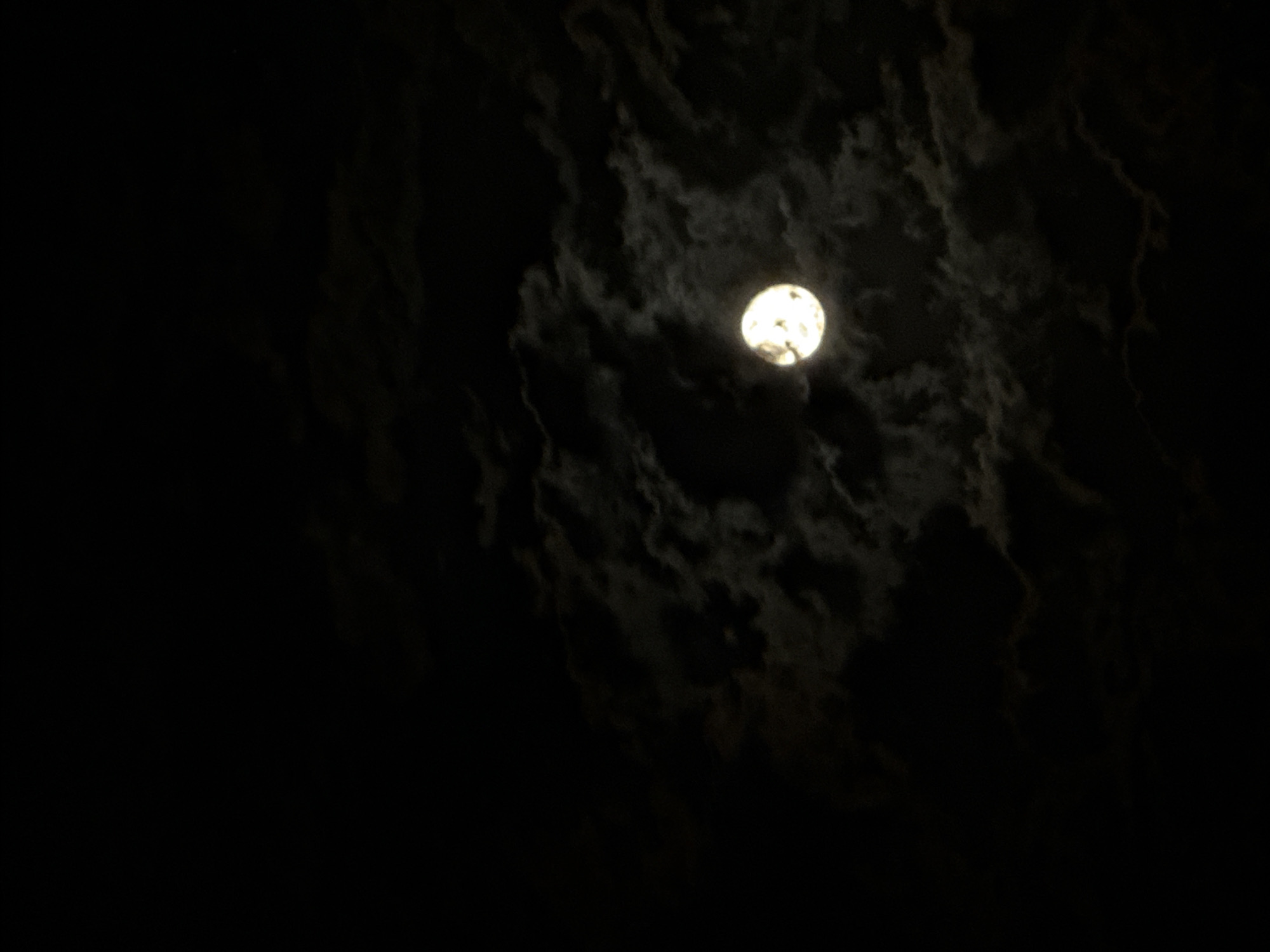 The image presents a nocturnal scene dominated by a bright, full or nearly full moon partially obscured by clouds. The moon, a luminous sphere of white and pale yellow, is positioned in the upper right quadrant of the frame, casting a soft glow that highlights the undulating shapes of the surrounding clouds. These clouds are dark, ranging from deep charcoal to lighter shades of grey where the moonlight diffuses through them, creating a textured and somewhat ethereal quality to the night sky. The overall background is a deep, unlit black, emphasizing the solitary brilliance of the moon and the shadowy forms of the clouds. There are no human subjects, man-made objects, or discernible terrestrial features. The activity depicted is the natural phenomenon of the moon shining through clouds at night. The time of day is clearly night, with the weather being cloudy. No specific location cues are visible beyond the atmospheric elements. There is no visible text in the image.