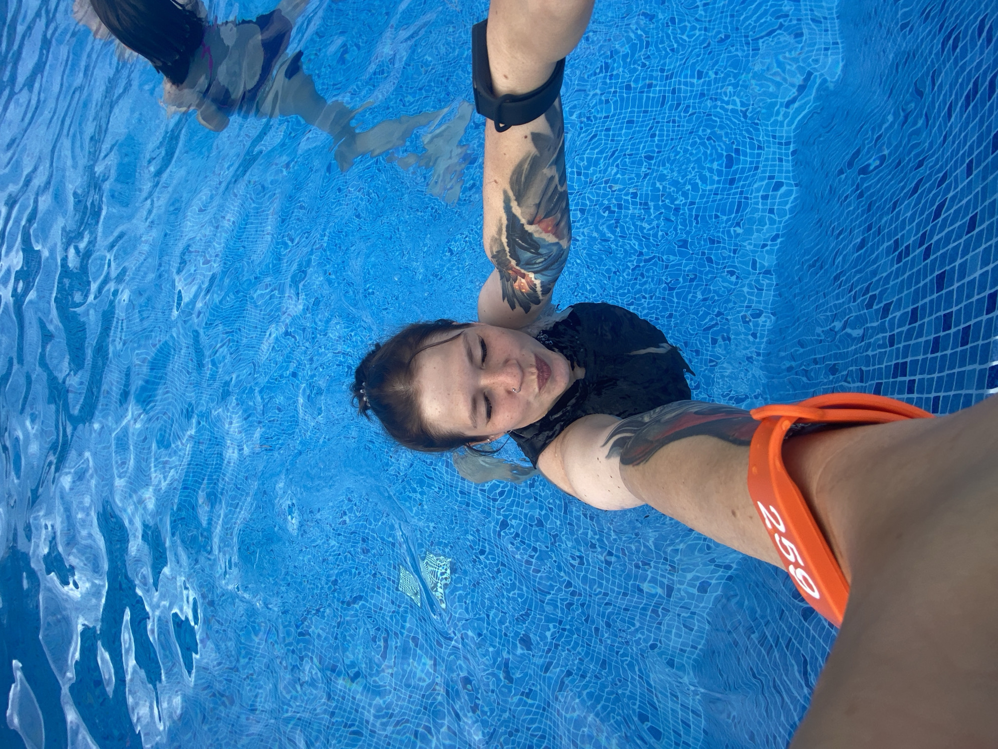 A first-person perspective selfie is captured in a blue swimming pool. A woman with her hair in a bun, wearing a black swimsuit, is posing for the camera with a playful expression. She has tattoos on her left arm and a nose ring. An orange wristband with the number "259" is visible on her right arm. The water is clear and shows the tiled bottom of the pool. Another person is partially visible in the background, further back in the pool. The setting appears to be an outdoor pool, given the natural lighting and the ripple patterns on the water's surface.