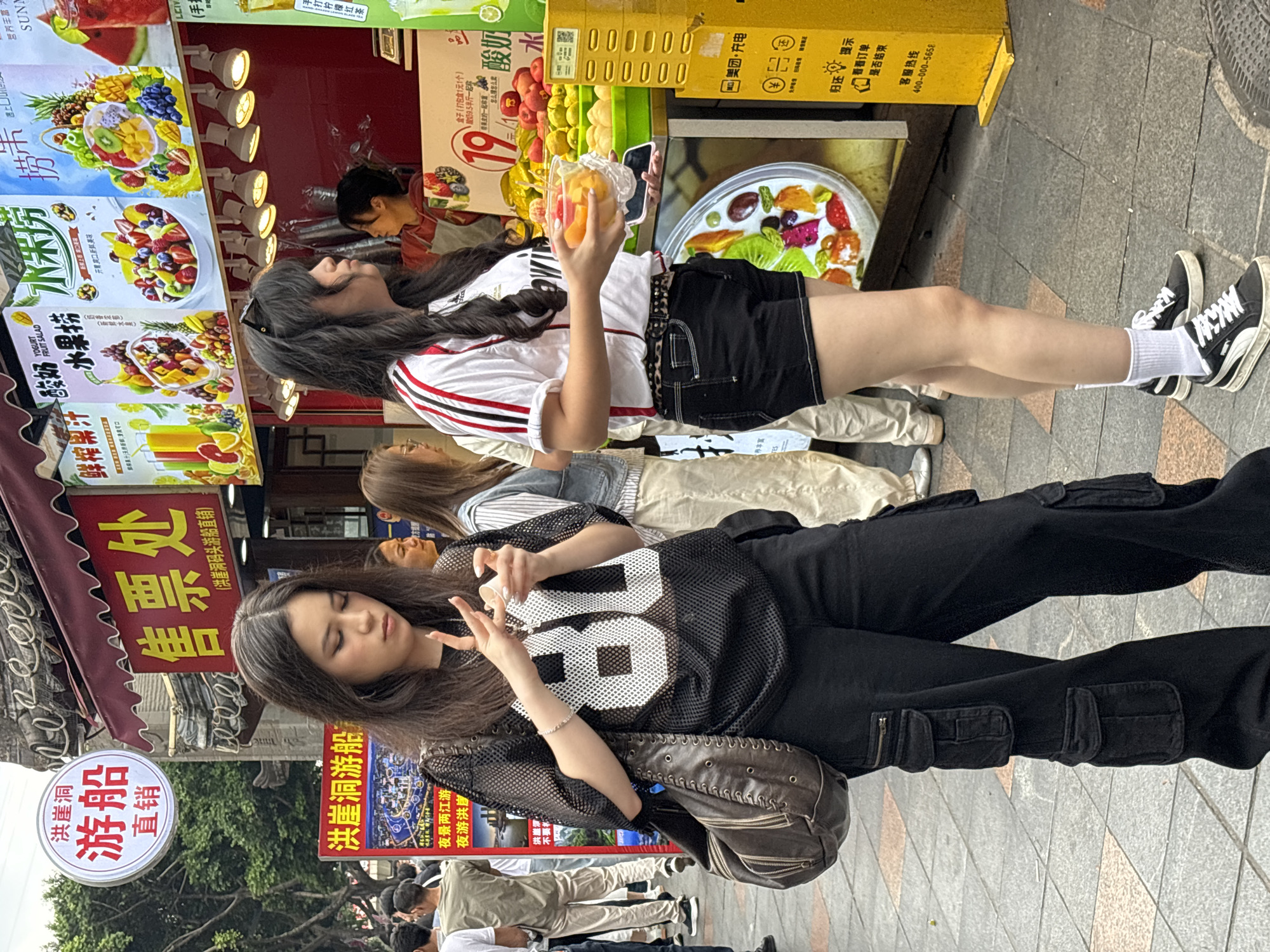 A bustling street scene in Yuzhong District, China, shows two young women standing in front of a fruit stand, surrounded by signs in Chinese. The woman in the foreground wears a black mesh jersey with the number 88, black cargo pants, and carries a brown leather bag, making a hand gesture. The other woman is wearing a white and red striped shirt and black shorts and is holding a clear container of fruit. In the background are signs for a “Hongyadong” cruise, advertising “Night views of the two rivers, night cruise of Hongyadong.” The fruit stand is adorned with colorful pictures of various fruit salads and juice combinations. The street is crowded with pedestrians.