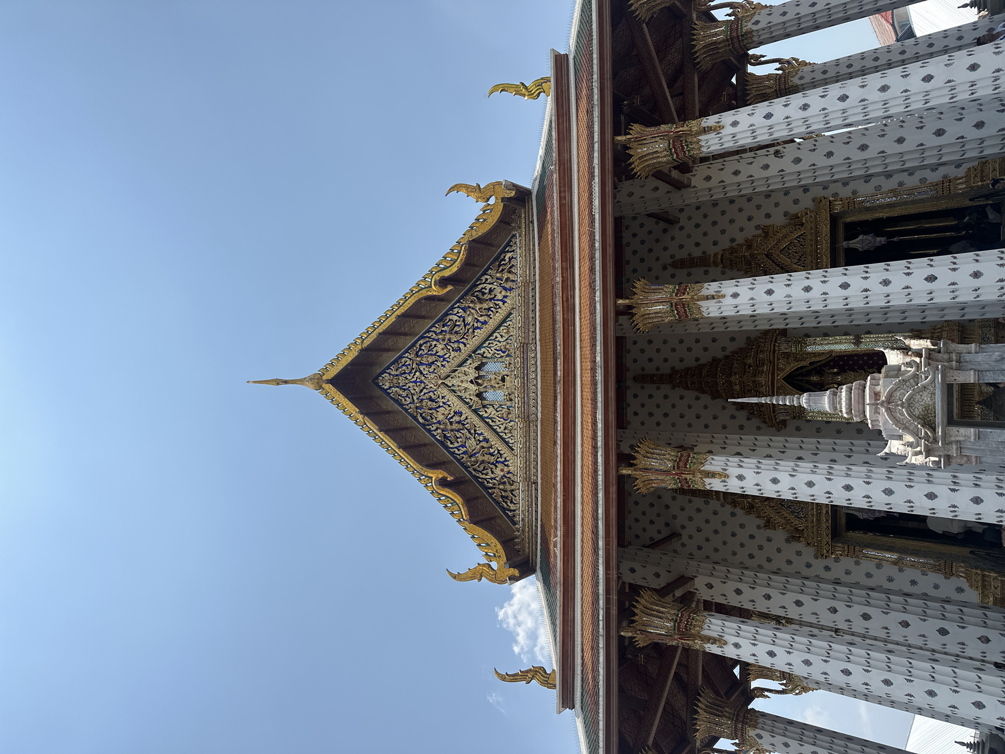 The image depicts a section of the Wat Arun Ratchawararam Ratchawaramahawihan, more commonly known as Wat Arun or the Temple of Dawn, in Bangkok, Thailand. It is an upward-angled shot of the temple's exterior architecture.
The image captures a portion of the temple's ornately decorated roof with intricate carvings and gold accents. The triangular gable above the columns is detailed with floral and geometric patterns. The roof is tiled in a series of earth tones, with decorative golden finials along the edges. Below the roofline, a row of white columns with decorative patterns are visible. The columns are also ornamented with gilded details at their bases and tops. The background consists of a clear, blue sky.