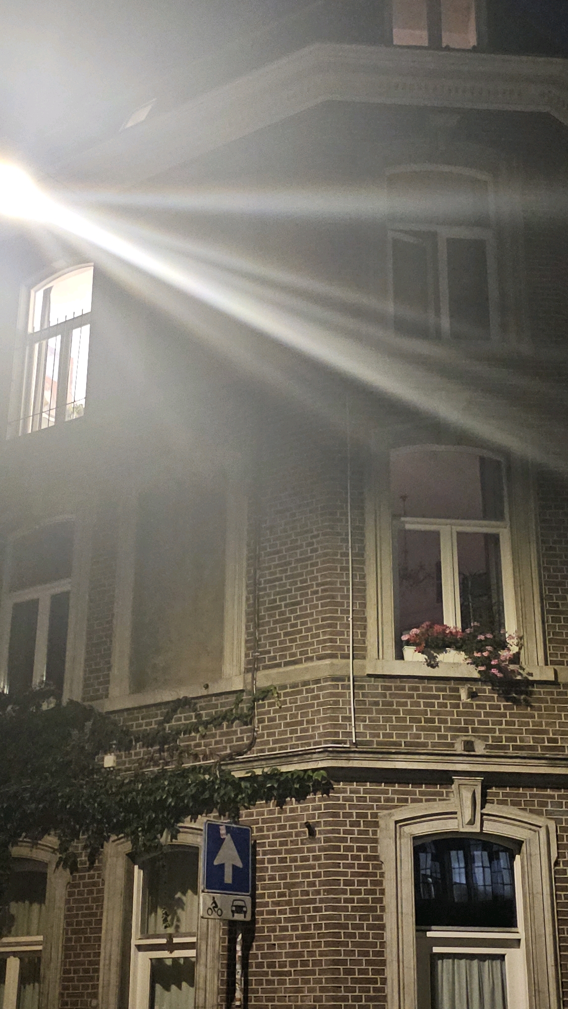 The image depicts a street-level view of a brick building at night, with strong beams of light cutting across the facade. The architecture suggests a European city, likely Maastricht, Netherlands, given the context. The building features multiple windows, some with curtains visible. One window on the second floor has a flower box filled with pink and red flowers. Climbing ivy is present on the lower sections of the building. A street sign indicating a one-way street for cars and bicycles is visible to the left of the building. The lighting suggests a strong, artificial light source, possibly a spotlight or streetlamp, creating dramatic rays. There are no people or animals visible in the image.