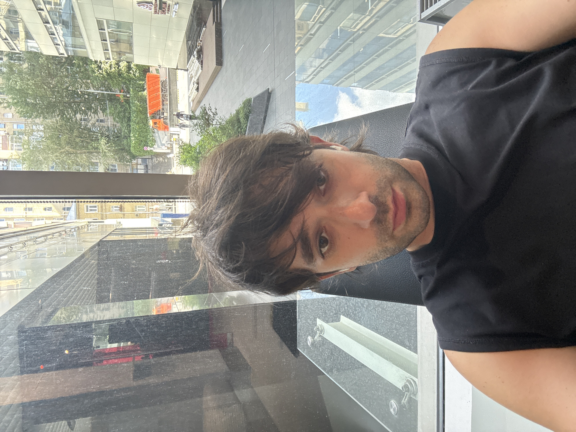 A young man with dark, disheveled hair is shown in a close-up selfie, looking directly at the camera. He is wearing a black sleeveless t-shirt. The background suggests an urban environment, possibly Moscow, Russia, with buildings and trees visible through a window or glass facade. Reflections in the glass indicate an outdoor setting with daylight. To the right of the man, a modern building with glass windows is visible, and further in the distance, an orange construction truck and a few pedestrians can be seen on a street. The overall scene appears to be taken from an indoor vantage point overlooking a city street.