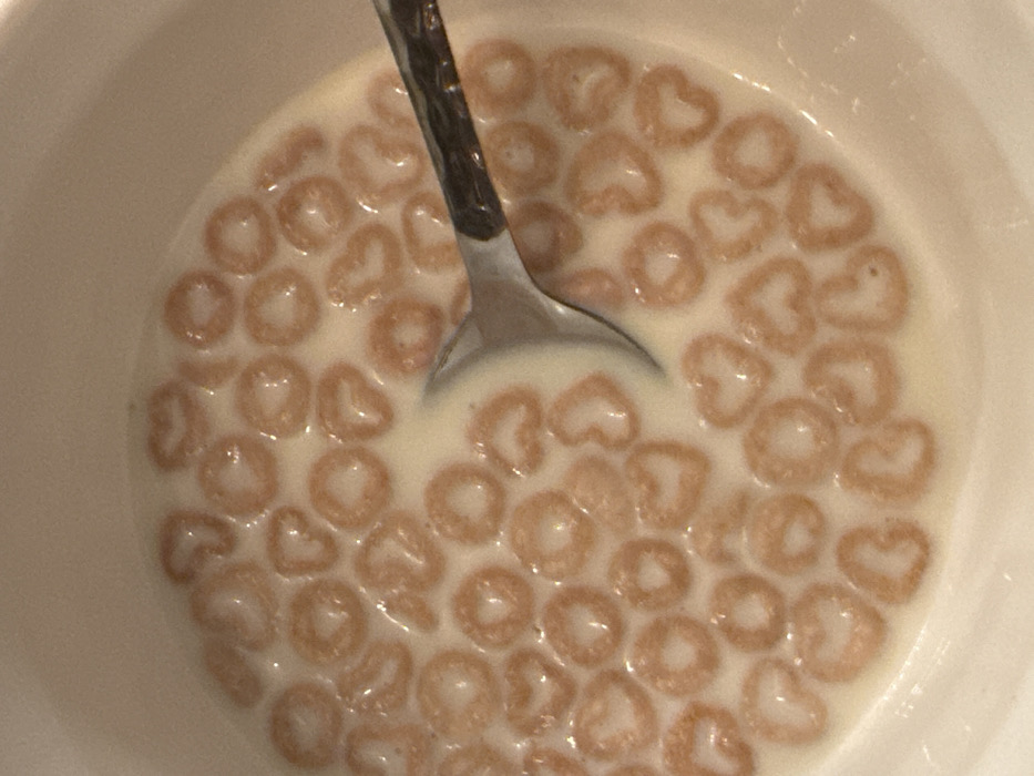 A top-down shot shows a white bowl filled with heart-shaped cereal and milk. A silver spoon rests in the center. The milk appears thin and lightly coats the cereal. The cereal is a light tan color, and numerous pieces are visible floating evenly in the milk. The bowl's rim is partially visible along the edges of the frame. The scene is lit softly, creating a gentle and inviting presentation.
