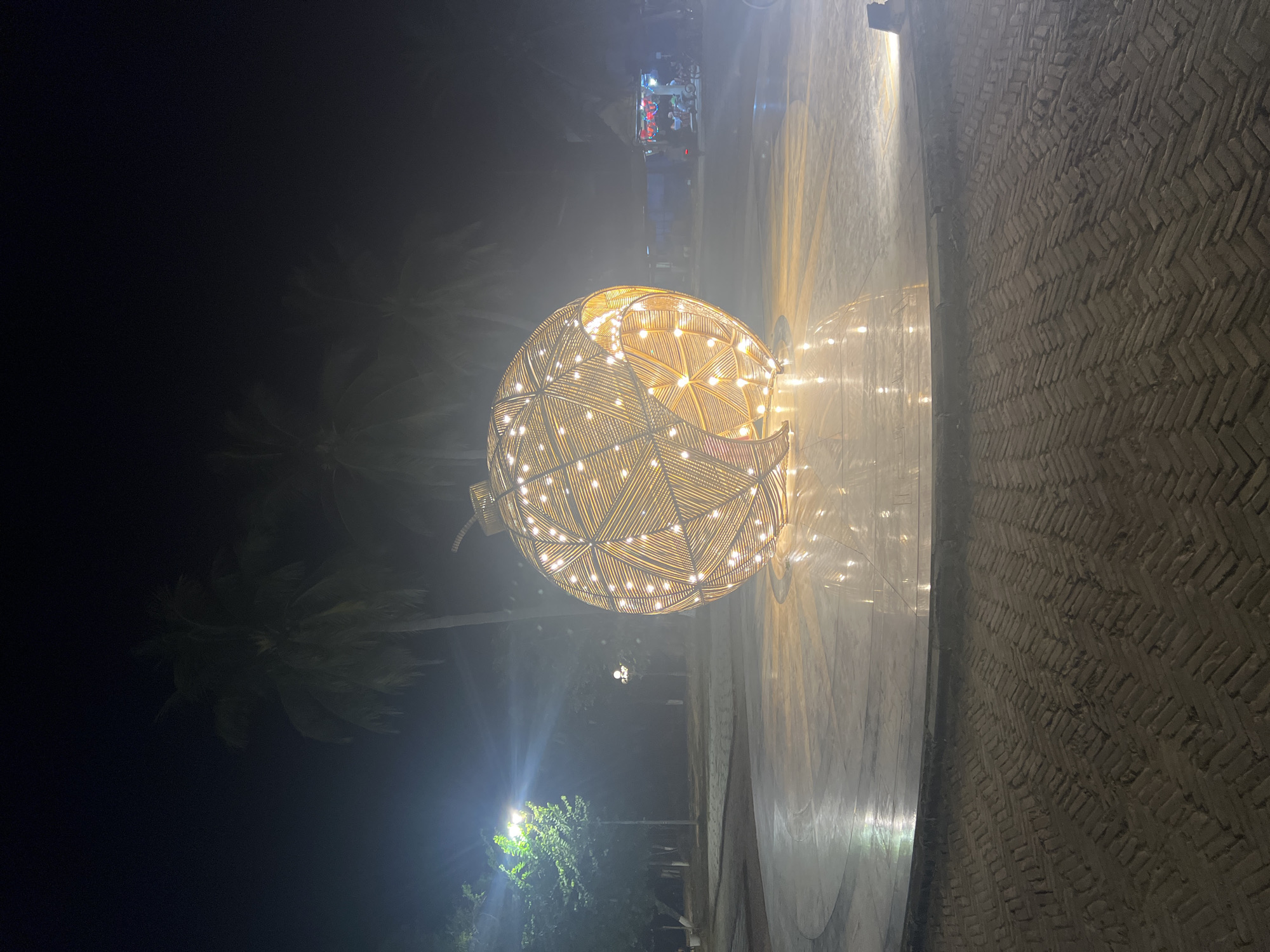 This media file captures an outdoor scene at night, illuminated by artificial lights. The primary subject is a large, spherical decorative light fixture, resembling a woven orb, adorned with numerous small, warm-toned lights. The orb is placed on a paved area, which transitions from a smooth, reflective surface on the left to a textured brick pattern on the right. The lighting from the orb casts a soft glow on the surrounding pavement and creates reflections.
In the background, palm trees are visible, silhouetted against the dark sky, suggesting a tropical or subtropical environment. There are also streetlights in the distance, one casting a bright green light on foliage and another providing a softer illumination. A hint of a structure or activity can be seen in the upper background, with some colorful lights suggesting a stall or a gathering.
The overall atmosphere is serene and festive, with the decorative lighting contributing to a pleasant ambiance. The time of day is clearly night, and the weather appears calm, with no indications of rain or strong winds, although some atmospheric haze or mist might be present due to the lighting. The location cues, such as palm trees and the overall architectural style of the paving, are consistent with a warm climate. No visible text is present in the media file.