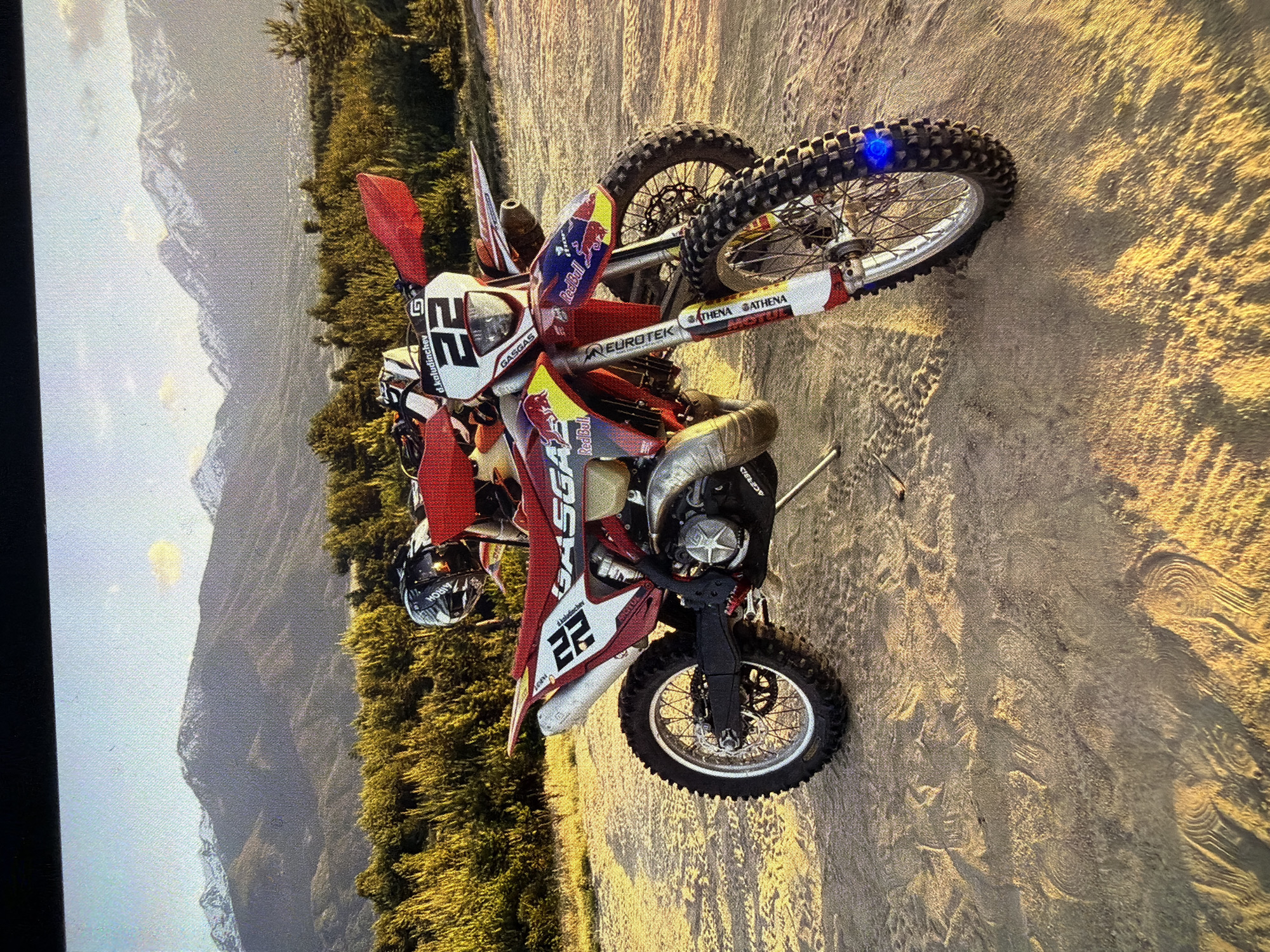 An action shot features a red and white GasGas motocross or enduro dirt bike, prominently displaying the number "22" on its front and side panels. The bike is parked on a sandy or dirt track, with clear tire impressions visible on the ground, suggesting recent off-road activity. The frame is adorned with multiple sponsor decals, including "Red Bull," "ACERBIS," "EURO TEK," "ATHENA," "Jinchen," and "d.kaludinchev." A black "AIROH" brand helmet rests on the ground near the front of the bike.
The setting appears to be a natural, rugged landscape. In the mid-ground, a dense forest of green trees stretches horizontally across the image. Beyond the treeline, a range of large, rugged mountains rises under a bright, possibly hazy, sky. The lighting, characterized by long shadows and a golden hue illuminating the ground and parts of the bike, indicates either early morning or late afternoon. The overall scene suggests a pause during an adventurous off-road ride in a mountainous region, consistent with the provided location context of Bania, Bulgaria.