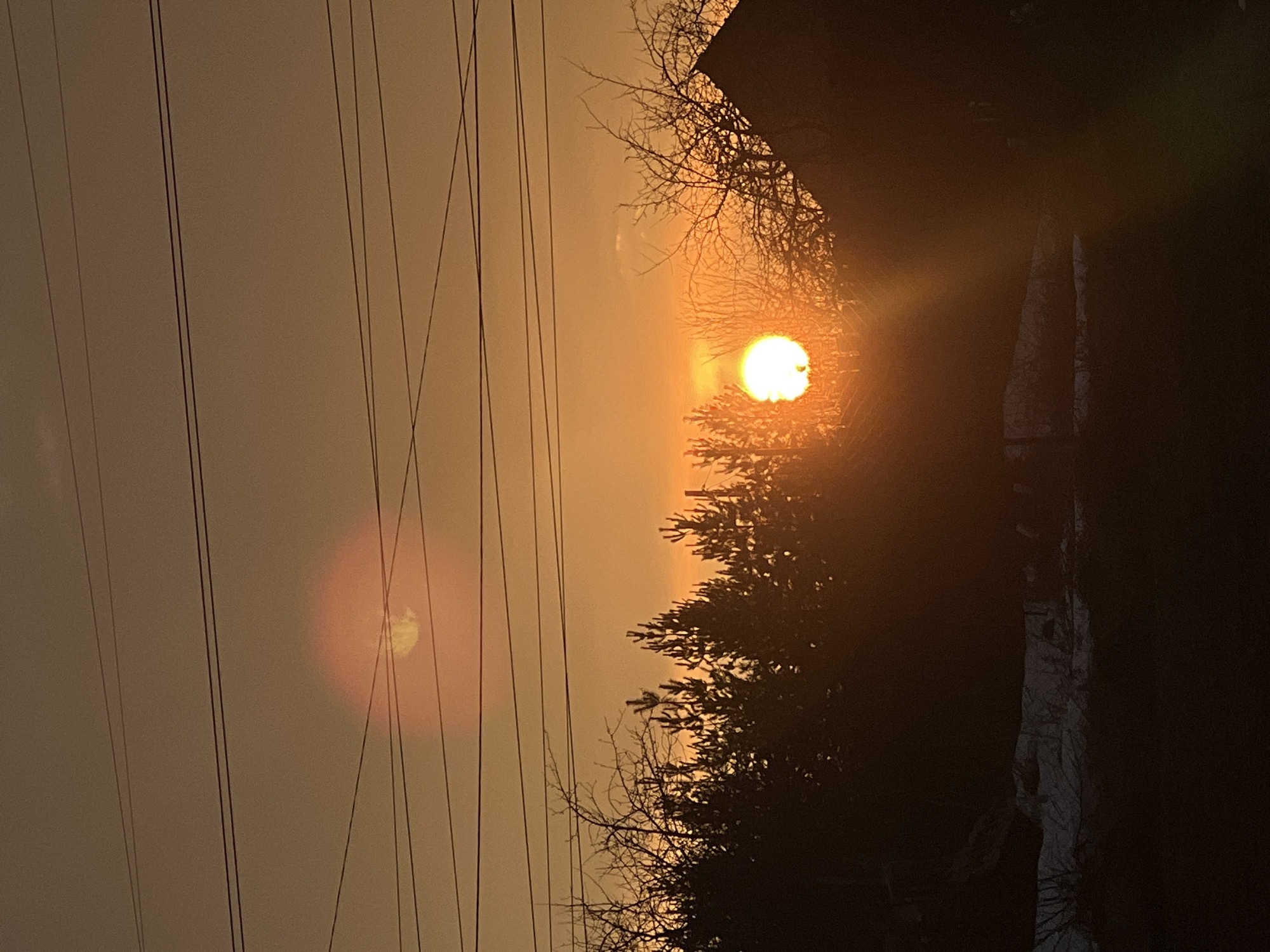 The image shows a view in the city of Chaikovsky, Russia, at sunset. Overhead power lines run vertically on the left. A bright, orange sun is partially obscured by trees in the center of the frame. To the right, there is a dark area which appears to be a building and some snow or ice. The sky is hazy with a pale orange tint. There is a sun halo visible on the left.