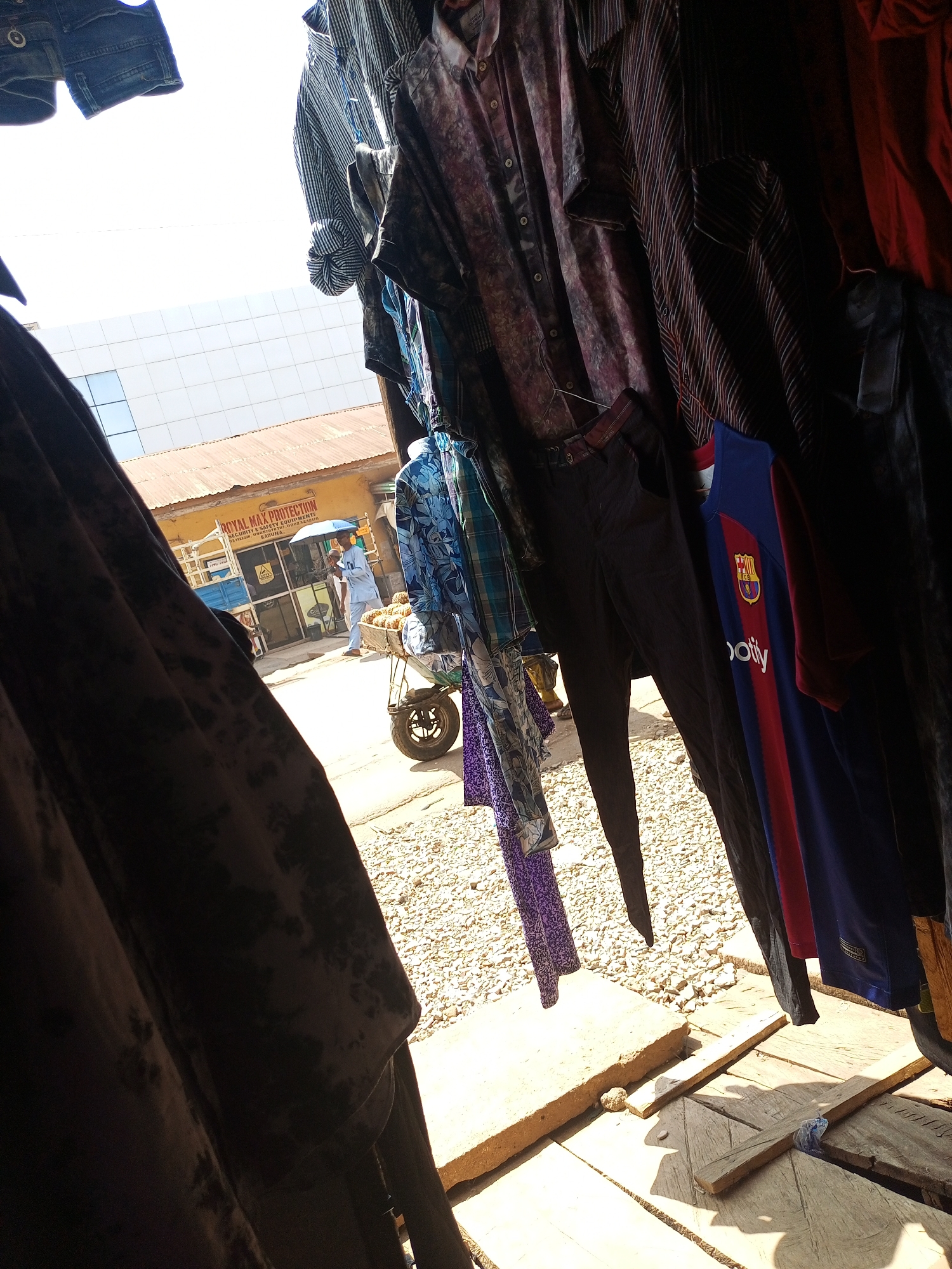 The image captures a vibrant street scene, viewed through an opening framed by hanging clothing, likely from a market stall or shop. In the immediate foreground, various garments are suspended, creating a dark border on the left and right sides. These include dark denim shorts at the top left, and on the right, several shirts of different patterns and colors, dark trousers, and a prominent blue and red FC Barcelona football jersey with the "Spotify" logo and club crest visible.
Beyond the clothes, the scene opens to a bright, sunlit outdoor area. The ground is unpaved, covered with light-colored gravel or small stones, with some wooden planks or pallets laid out. A man, dressed in light-colored traditional attire, is walking away from the camera, holding a light blue umbrella for shade. He is positioned next to a handcart or wheelbarrow, laden with what appears to be a pile of pineapples or similar fruit.
In the mid-ground, a single-story yellow building with a corrugated metal roof is visible. A sign on this building reads "ROYAL MAX PROTECTION" and "SECURITY & SAFETY EQUIPMENTS," with "KADUNA" partially visible below. Above this building, a modern, multi-story structure with a light-colored, possibly tiled or paneled facade reflects the bright sky. The sky itself is very bright, almost white, indicating strong midday sun. The harsh sunlight creates sharp shadows on the ground.
The overall setting suggests a bustling market or commercial area in Maiburiji, Nigeria, characterized by outdoor vendors and a mix of traditional and modern architecture.