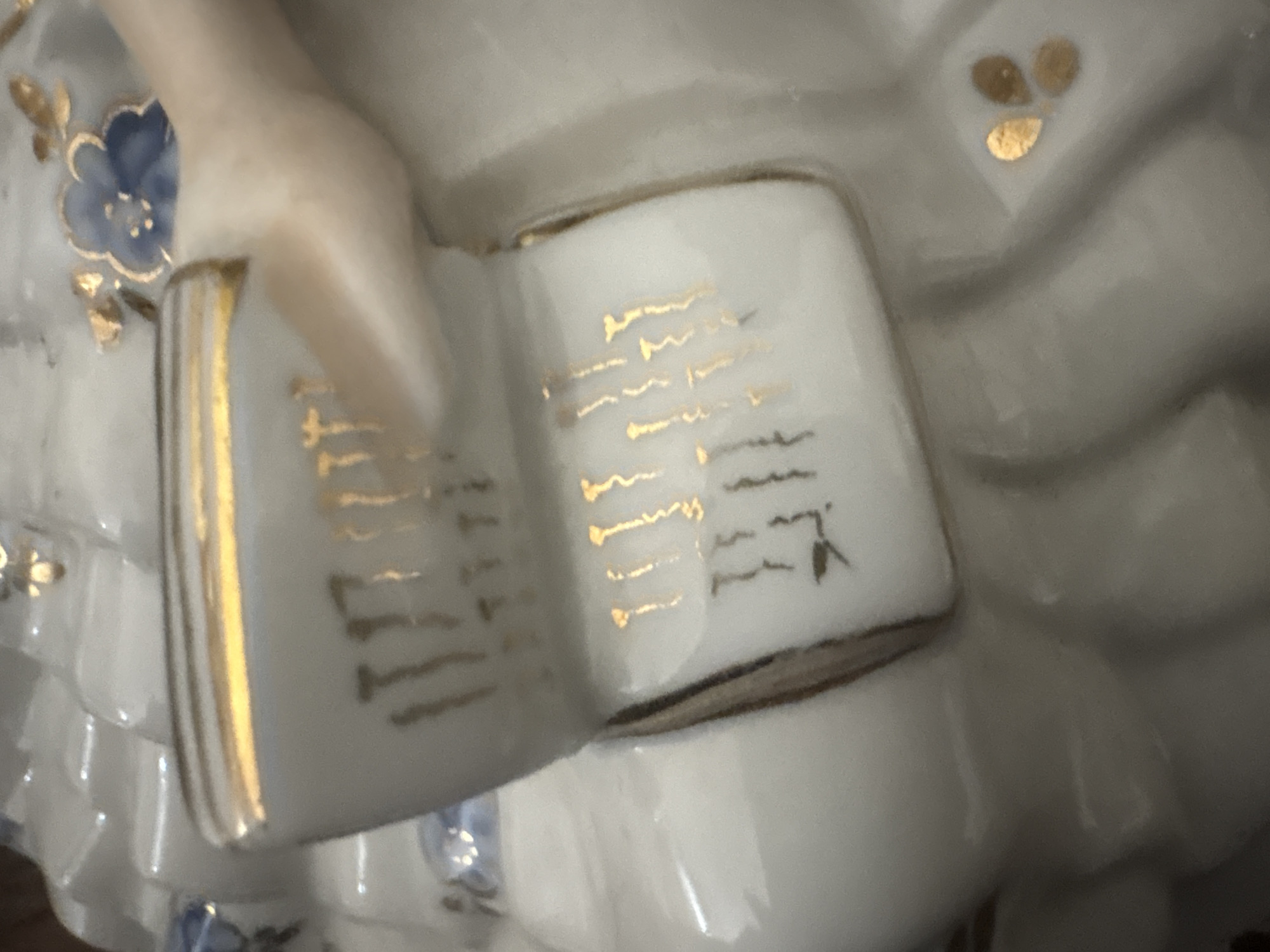 This is a close-up, high-angle, indoor shot of a porcelain figurine of a woman holding an open book. The figurine is white with gold accents and blue floral details. The book's pages are depicted with gold lettering that appears to be a decorative pattern rather than legible text. The woman's right hand and arm are visible, resting on the book. The background is blurred and indistinct, suggesting a shallow depth of field. No specific location in Georgiyevsk, Russia, can be identified from the image.