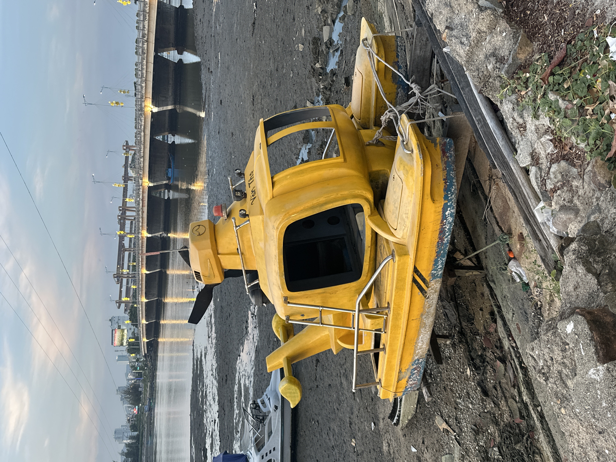 A yellow, helicopter-shaped boat is resting on a muddy shore in the city of Nam Nha Trang Ward, Vietnam. The boat has a rotor on top, windows, and two floats on the sides. There is a ladder leading up to the boat, and metal railings around the deck. In the background, a bridge spans a body of water, with buildings and power lines visible in the distance. The sky is overcast with scattered clouds. The shore is covered in mud, debris, and some sparse vegetation. Another white boat is visible to the left of the yellow boat. The scene appears to be a low tide situation.