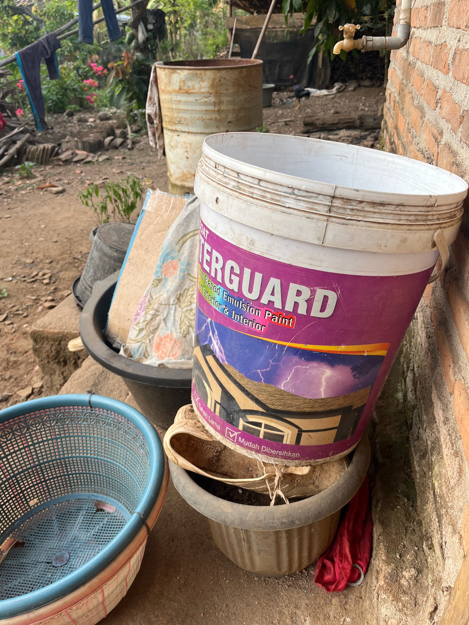 The image shows an outdoor scene in Kebumen, Indonesia, with a focus on discarded items. A large, white plastic bucket with a purple label for "WATERGUARD" water-based emulsion paint is prominently featured, leaning against a red brick wall. The bucket is stacked on top of other containers, including a dark-colored pot containing dirt and a shoe. To the left of the paint bucket, a blue plastic basket is visible. In the background, laundry hangs on a line, and a rusty metal barrel stands next to some pink flowers. A water tap is attached to the brick wall. The ground is dirt and scattered with debris. The scene appears to be in a residential or working area, possibly a backyard or an alleyway. There are no people directly visible in the frame.