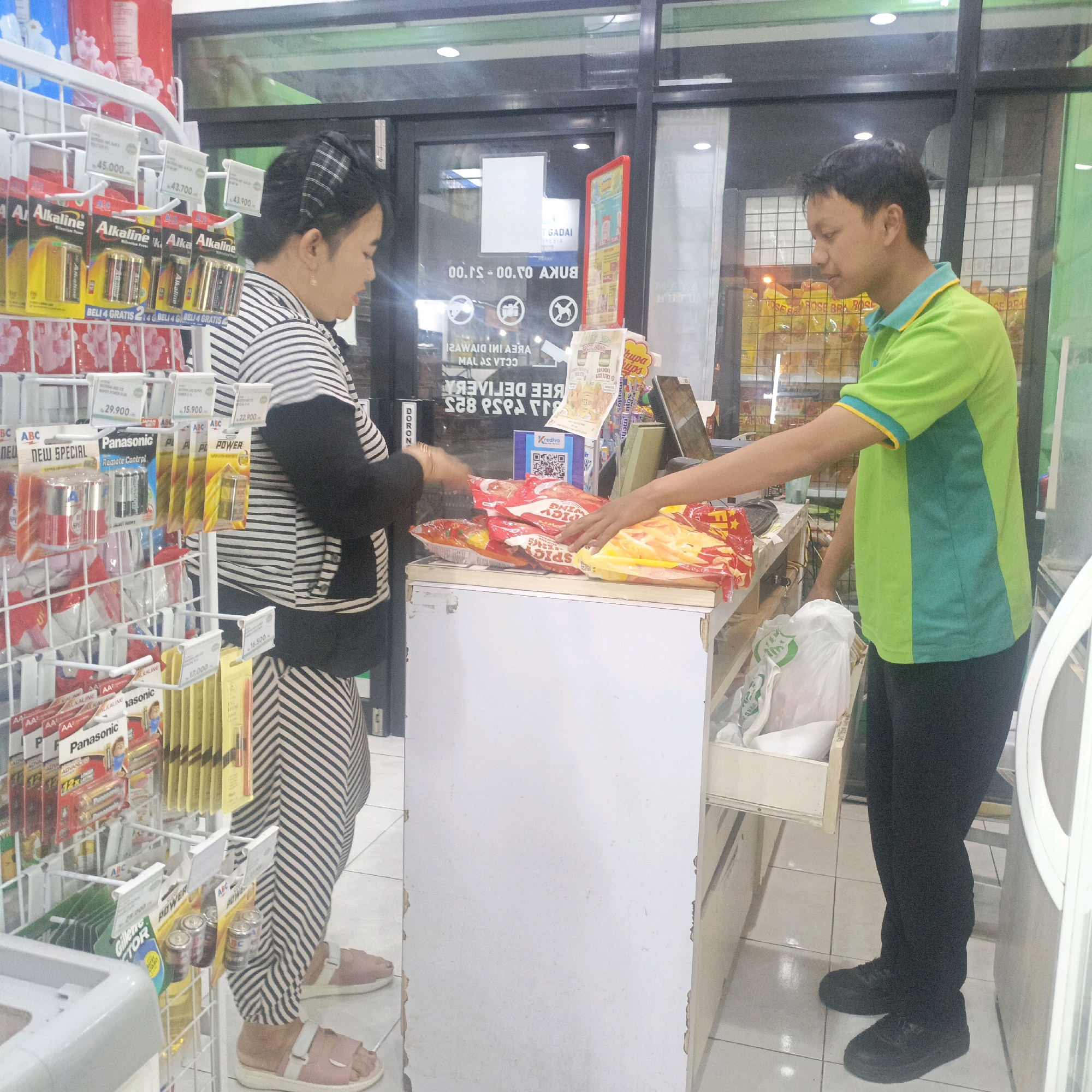 The image shows a retail store interior, likely a convenience store or minimarket, in Salatiga, Indonesia. A woman in a black and white striped outfit is standing in front of a display rack filled with batteries and other small items. She is reaching out towards a counter where a man in a green and blue polo shirt and black pants is standing. The man is reaching for a bag of snacks or chips on the counter. There are various products visible on shelves, including batteries, snacks, and drinks. A price tag with "45.000" is visible on the battery display. A digital display or screen is also visible in the background, showing information about store hours and services like "GADAI INDONESIA" (Indonesia Pawnshop). The overall environment suggests a typical commercial setting.
