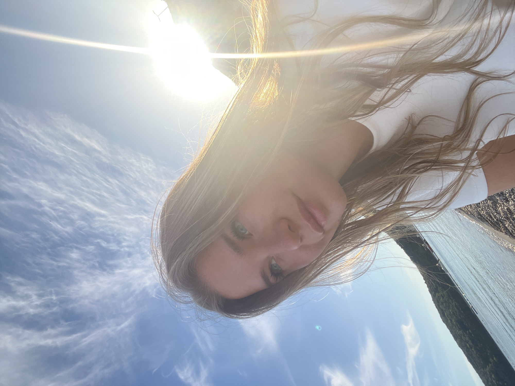 A young woman with long, light brown or blonde hair and light blue/green eyes is the central subject of this outdoor photograph. Her face occupies the upper-center of the frame, with her gaze directed downwards towards the camera, suggesting a selfie or a shot taken from a low angle. She wears a white top, parts of which are visible, and her hair flows around her head.
The setting is an extremely bright, sunny day. A brilliant sun flare emanates from the upper left corner, casting a strong, warm light across the scene and creating luminous highlights in her hair. The sky is a clear, vibrant blue, adorned with delicate, wispy white cirrus clouds that stretch across the expanse. To the right and slightly below the woman, a vast body of water is visible, possibly a lake or a wide river. Across the water, a distant landmass covered in dark green trees or foliage forms the opposite bank or shoreline. The immediate foreground near the water appears to be a rocky or pebbly surface.
The image conveys a sense of serene, sunny weather in a natural landscape. The strong sunlight and clear sky indicate it is likely midday or early afternoon. The location context of Prijevor, Montenegro, suggests this could be a lake or river within the country's mountainous terrain. No visible text is present in the image.