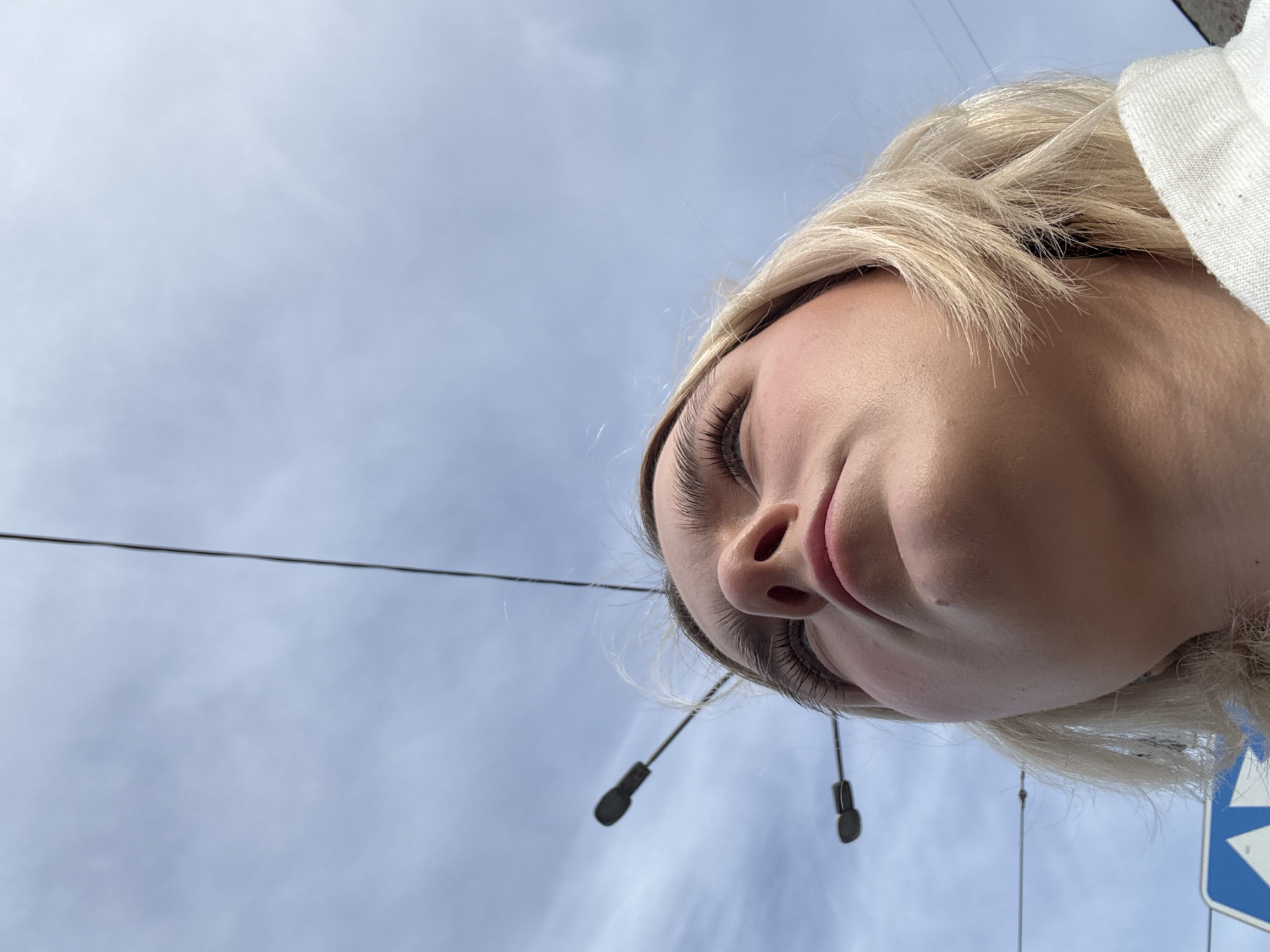 A low-angle shot features a person with blonde hair, facing downwards. The background is a light blue sky with wispy clouds and power lines. A white garment is visible at the top right corner. The person is wearing in-ear headphones. The scene is likely located in a city, as indicated by the presence of power lines and what appears to be a sign on the bottom right corner. While there are objects in the photo that hint to the location being the city of Vsevolozhsk, Russia it is not possible to verify that with the information in the image.