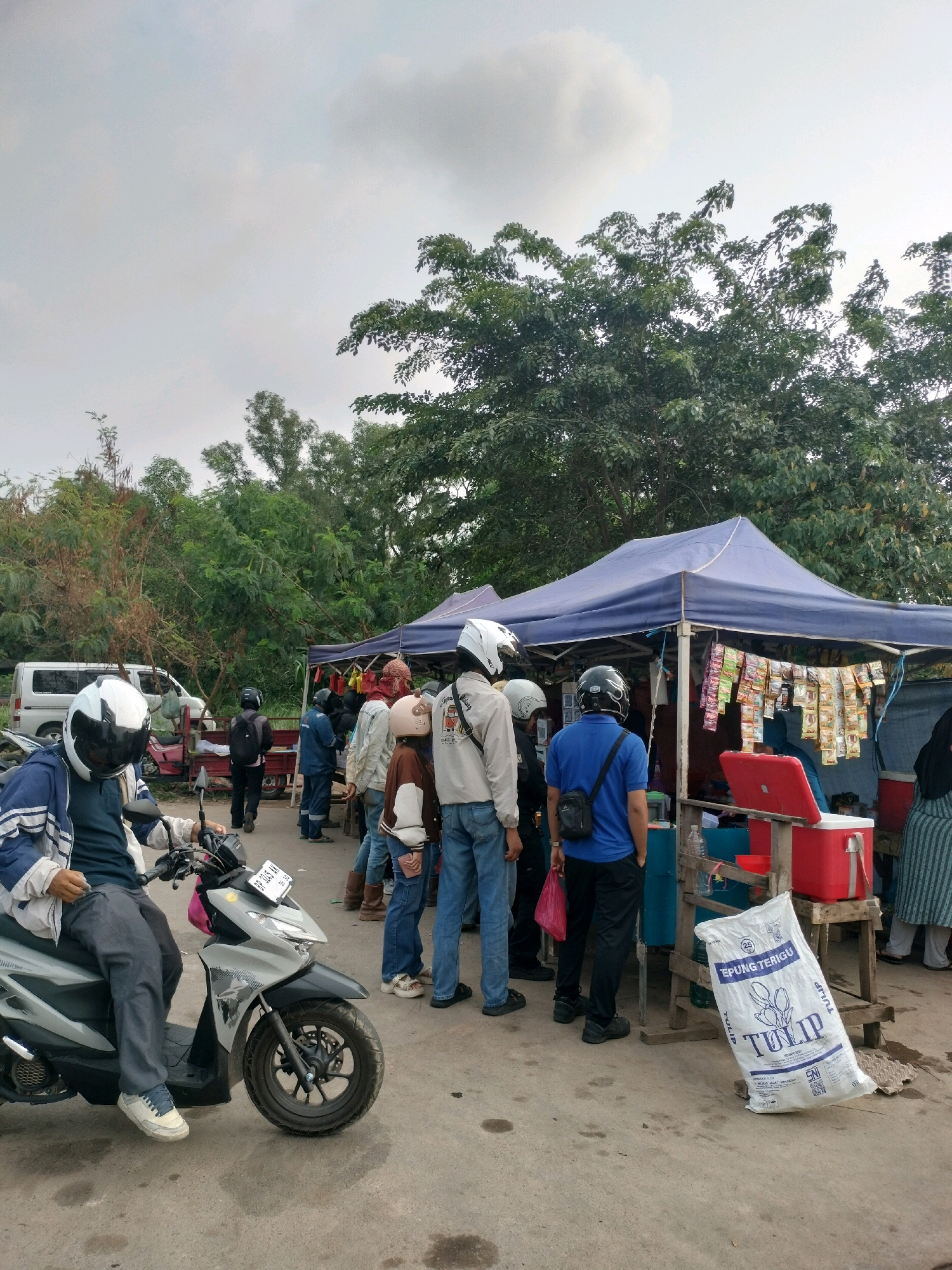 The image depicts an outdoor scene with several people gathered around a temporary stall under a blue tarp. The stall appears to be selling various items, with packets of snacks or goods hanging from the front. A large white bag with "TULIP" written on it is propped against the stall. In the foreground, a person wearing a white helmet is seated on a grey scooter. Other individuals, some also wearing helmets, are standing and interacting near the stall. Behind the stall, there are trees and vegetation, suggesting a semi-rural or roadside setting. A white van is visible in the background to the left. The overall atmosphere suggests a casual gathering or marketplace. This scene could be located in a peri-urban area of Batam, Indonesia, perhaps near a road or a local gathering spot.