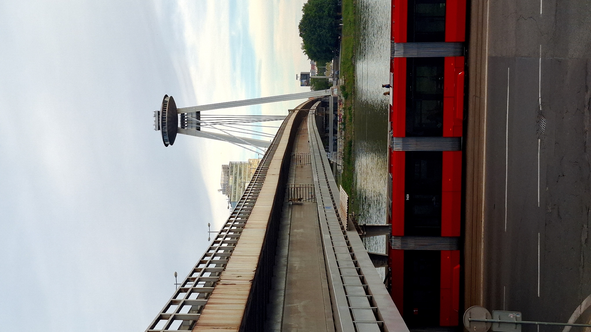 This image captures an urban landscape, likely in Bratislava, Slovakia, centered around the iconic Most SNP (UFO Bridge). The perspective is from the side of the bridge, looking along its length.
The most prominent subject is the **UFO Bridge** on the left, a cable-stayed bridge featuring a distinctive, saucer-shaped observation tower (the UFO Tower) at its top. The bridge's main pylon and cables are white, contrasting with the light, overcast sky. A pedestrian or maintenance walkway, made of concrete and metal railings, runs along the bridge structure, showing some signs of rust on its upper parts.
Parallel to the bridge on the right is a **river**, which is the Danube. Its surface reflects the sky and surrounding elements, with subtle ripples visible. Immediately adjacent to the river on the far right is a **road**, made of dark asphalt with white lane markings. Cracks are visible in the road surface.
A **red tram or articulated bus** with dark windows and silver corrugated sections is visible on the road. It appears to be a modern vehicle, and the number "7459" is discernible on its side. Two **people** are standing on the riverbank, looking towards the river or bridge, partially obscured by the red vehicle.
In the background, beyond the river, are green trees and various urban buildings. One building has the word "FOOD" partially visible. The sky is overcast or partly cloudy, indicating either an early morning, late afternoon, or generally cloudy day. The overall lighting is soft, not direct harsh sunlight. Street lights are present on the bridge walkway, and small road signs are visible at the bottom right of the frame.