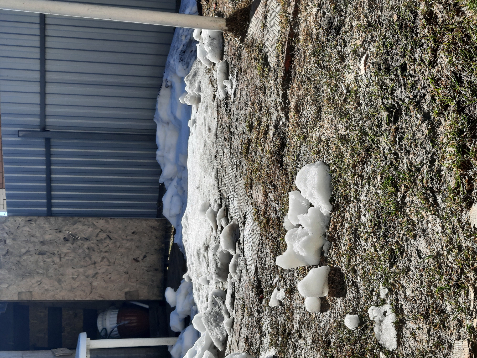 The image shows a patch of ground with melting snow in Krasnogorskoye, Russia. The area transitions from a concentration of white snow near a metallic structure and a wooden wall to a mix of brown grass and melting snow patches. The background includes a metal shed-like structure and a wooden surface. No people are visible in the image. The scene suggests the late stages of winter or early spring.