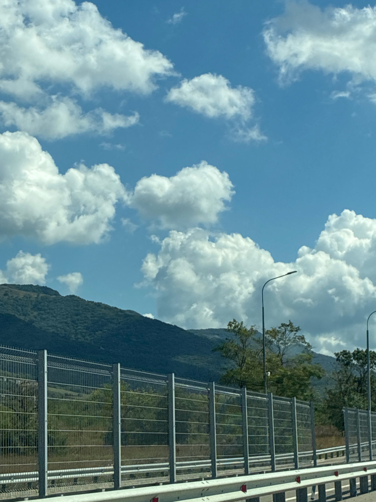 The image captures a view of a highway with a metal fence and a mountain in the background. The sky is filled with fluffy white clouds against a bright blue backdrop. There are streetlights visible along the highway, and trees are scattered on the side of the mountain. The mountain itself is covered with dense green vegetation, suggesting it is a natural landscape. The fence appears to be a sound barrier, common along major roads. The scene depicts a typical roadside view with natural elements and man-made infrastructure. There are no people or animals visible in the image. The location is identified as the city of Mar'ina Roshcha, Russia.
