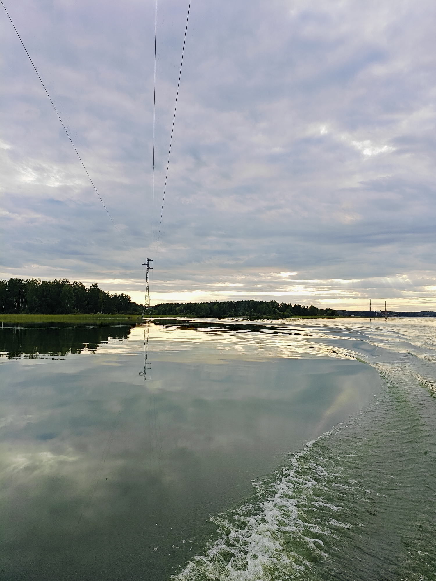 The image depicts a serene lake scene with a power line tower in the distance. The sky is filled with dramatic clouds, with rays of sunlight breaking through. The water is calm and reflective, mirroring the sky and the surrounding trees. The wake of a boat is visible in the foreground, suggesting movement and a journey across the water. The power line stretches across the sky, connecting to the tower, which stands tall amidst the natural landscape. In the far distance, two smokestacks rise above the treeline, hinting at industrial activity nearby. The overall impression is one of tranquil beauty, with a subtle juxtaposition of nature and human infrastructure. The scene is likely located on the Kama River or one of its tributaries near the city of Chaykovsky, Russia, given the prevalence of power lines and industrial elements in the region.